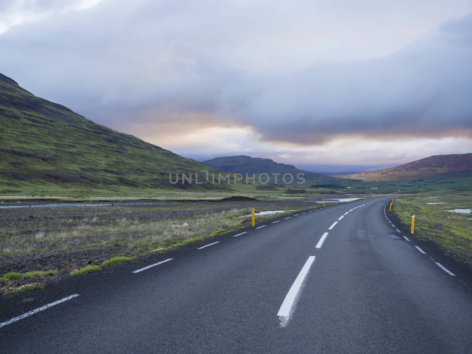 Asphalt road curve through empty northern landscape with green grass colorful hills, water puddles and sunset dramatic sky, way to the mountains in Iceland western highlands, copy space by Henkeova