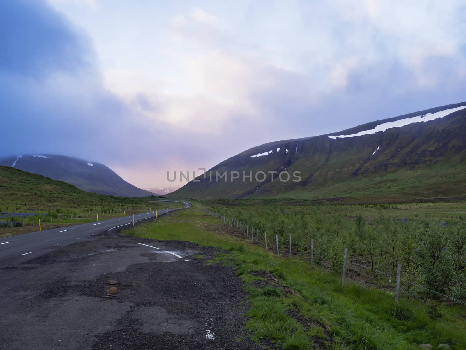 Asphalt road curve through empty northern landscape with green grass colorful hills and sunset dramatic sky, way to the mountains in Iceland western highlands, copy space