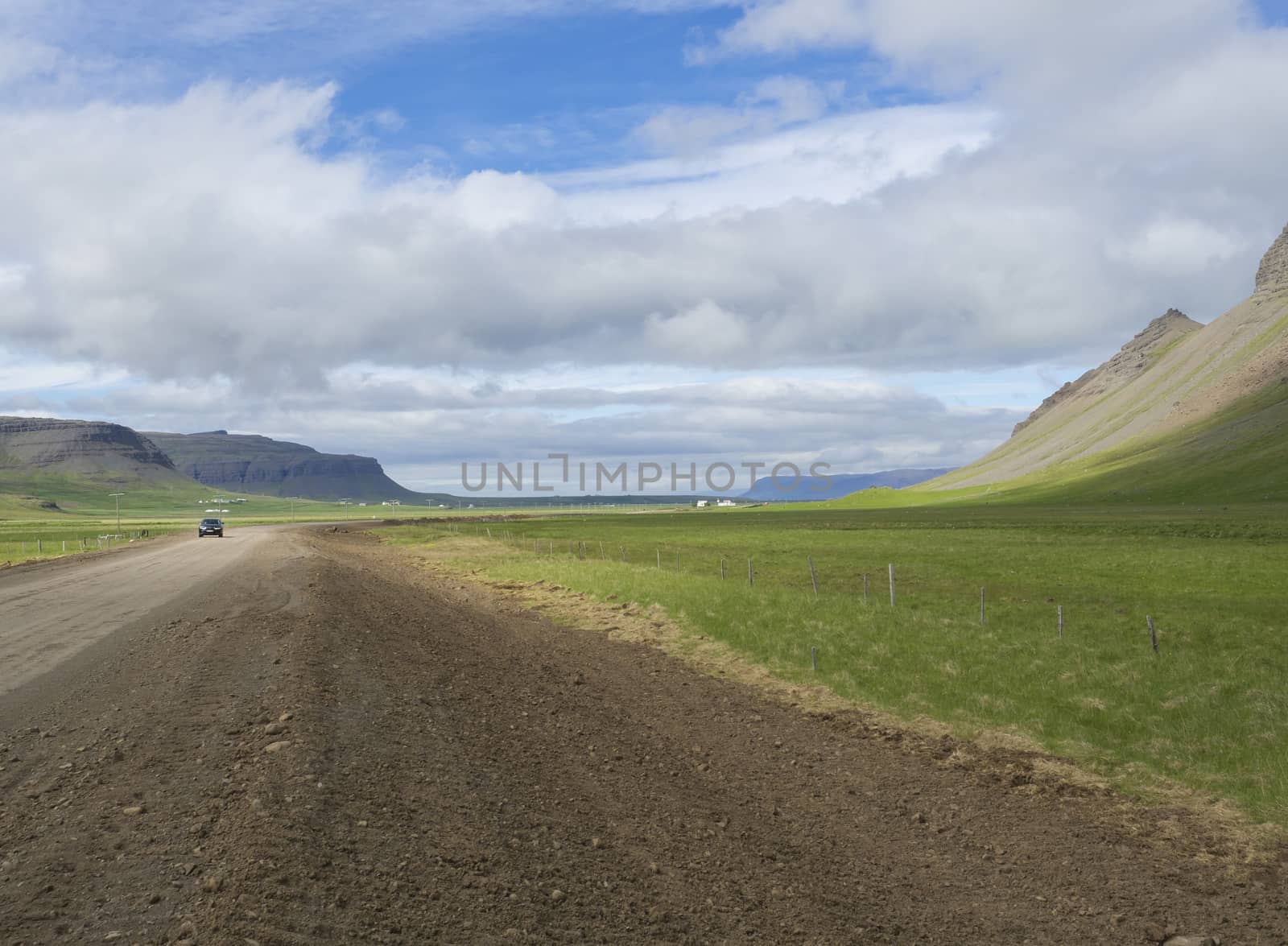 small black car driving on dirty road through valley in rural northern landscape with green grass and volcanic hills, abd farm houses, blue sky white clouds, summer in Iceland west fjords, copy space