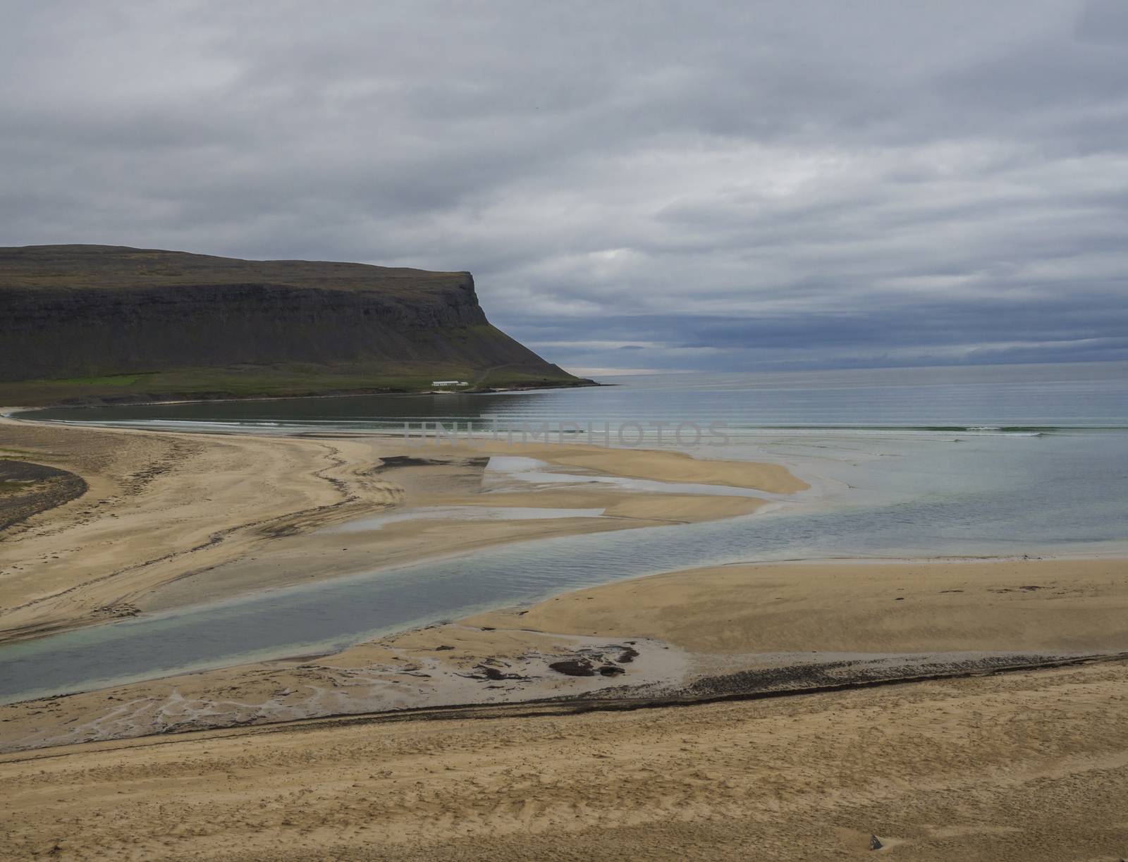 view on pink beige sand beach and green grass and stone shore in iceland west fjords with green cliff and ocean, river delta and moody sky background
