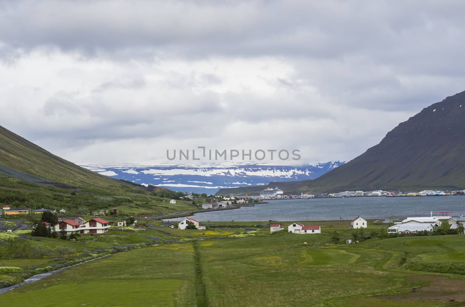 Beautiful view on Isafjordur city, capital of west fjords of Iceland in summer with red houses, harbor with ships and yachts, green grass and farm landscape, blue snow covered mountains background