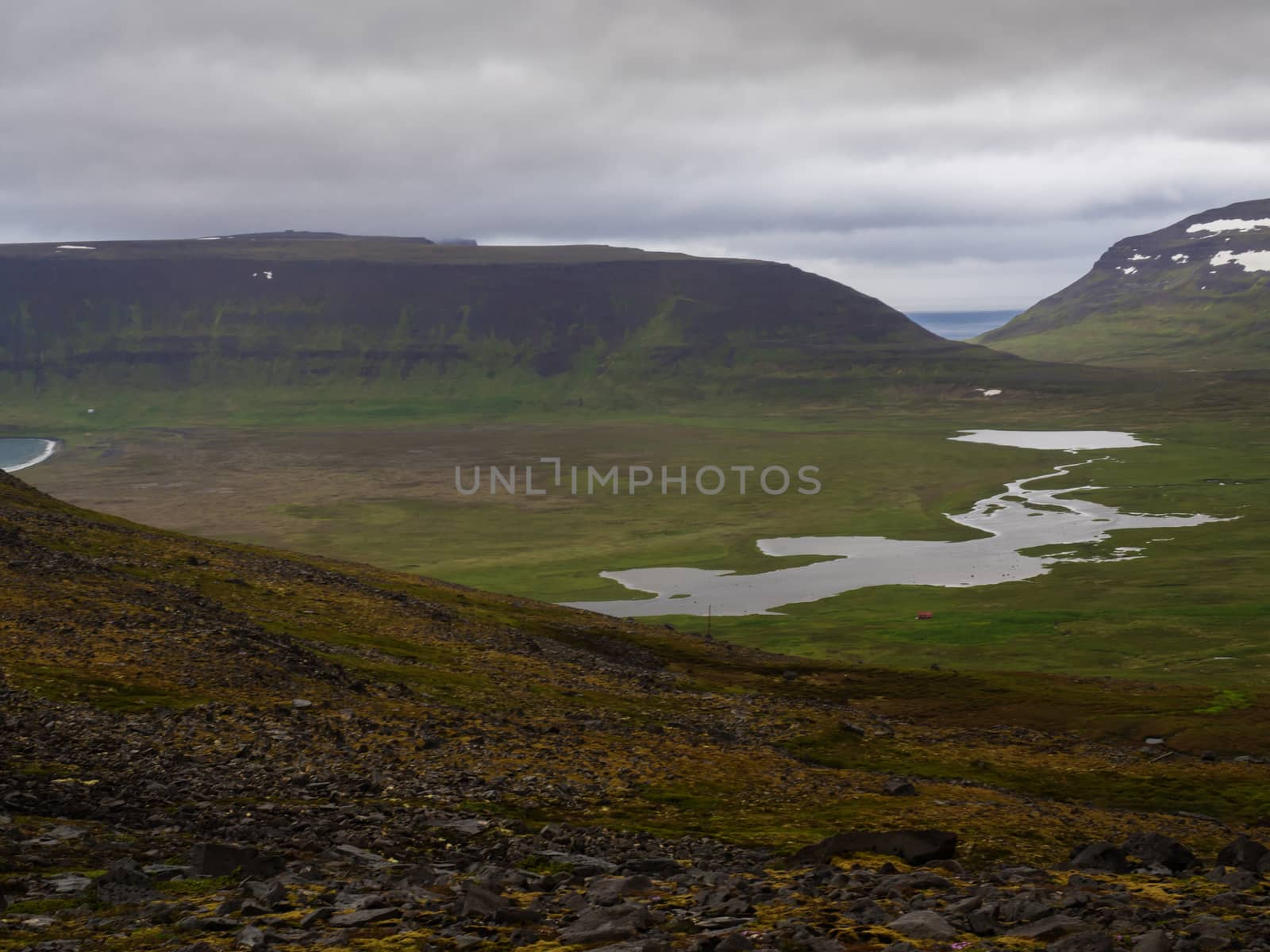 Northern summer landscape, beautiful snow covered cliffs and fljotsvatn lake in Fljotavik cove in Hornstrandir, west fjords, Iceland, with river stream, green grass meadow, moody sky background