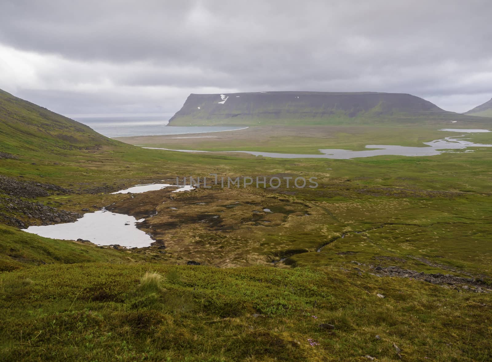 Northern summer landscape, View on beautiful cliffs in adalvik cove in west fjords nature reserve Hornstrandir in Iceland, with river stream delta, sand beach and sea, snow patched green meadow and dark clouds background