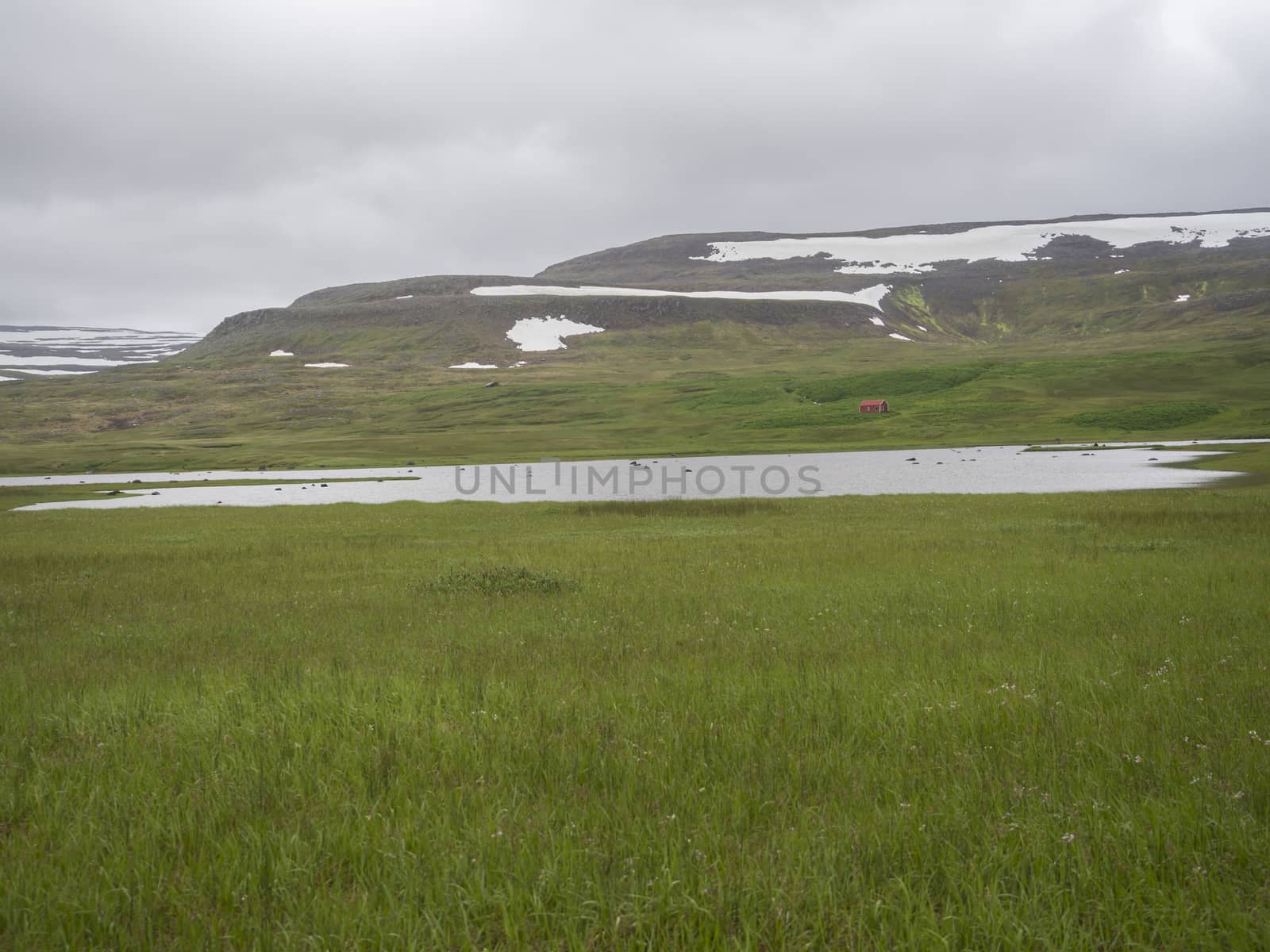 Abandoned red farm house in adalvik cove in west fjords nature reserve Hornstrandir in Iceland, with river stream, snow patched hills, green grass meadow and dark clouds background.