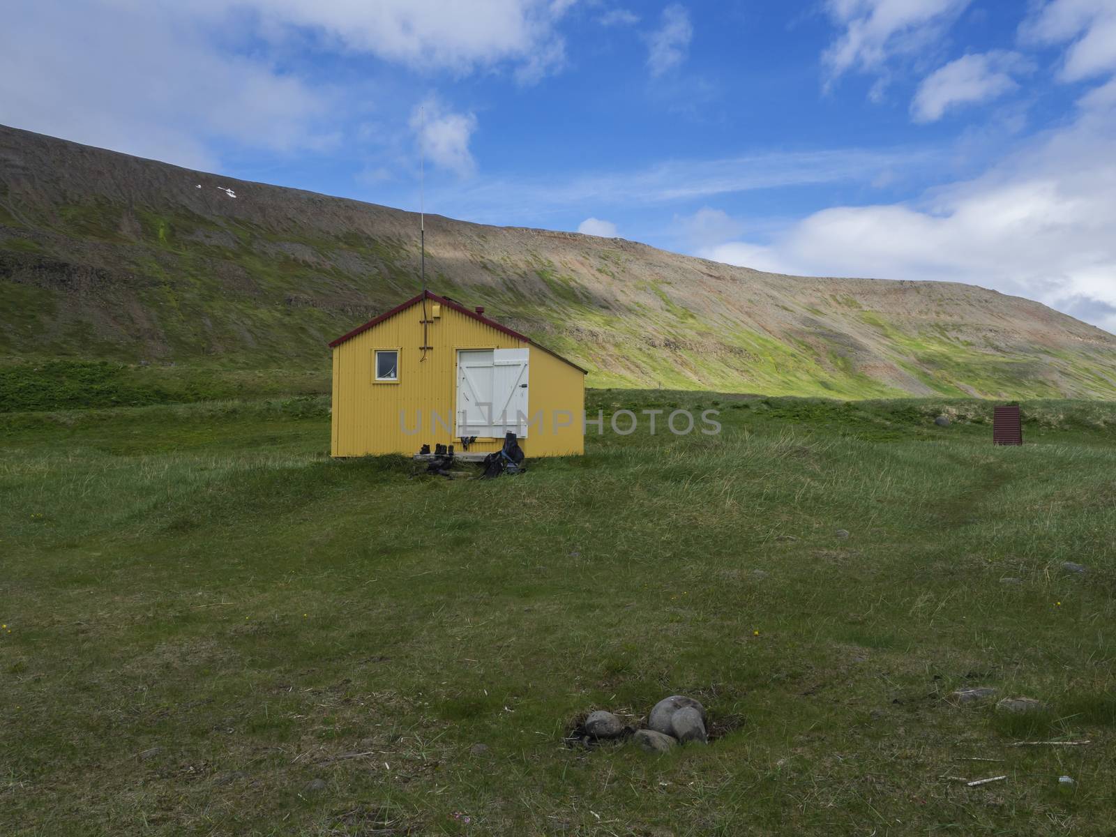 View on latrar camp site in adalvik cove with yellow emergency shelter cabin in west fjords nature reserve Hornstrandir in Iceland, with green grass meadow, hills and blue clouds background