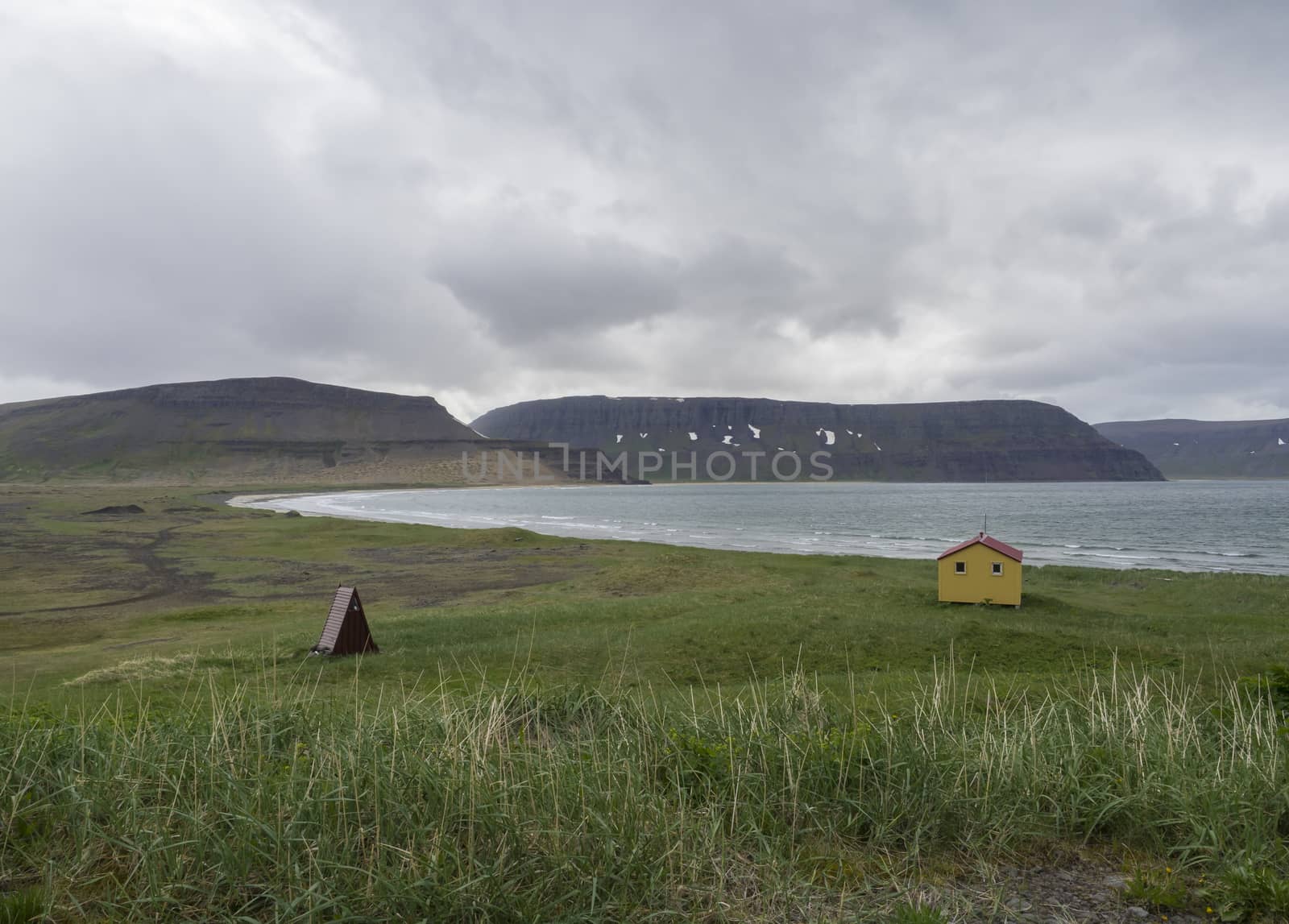 View on latrar camp site in adalvik cove with yellow emergency shelter cabin in west fjords nature reserve Hornstrandir in Iceland, with green grass meadow, stone beach, ocean, snow patched hills and dark clouds background by Henkeova