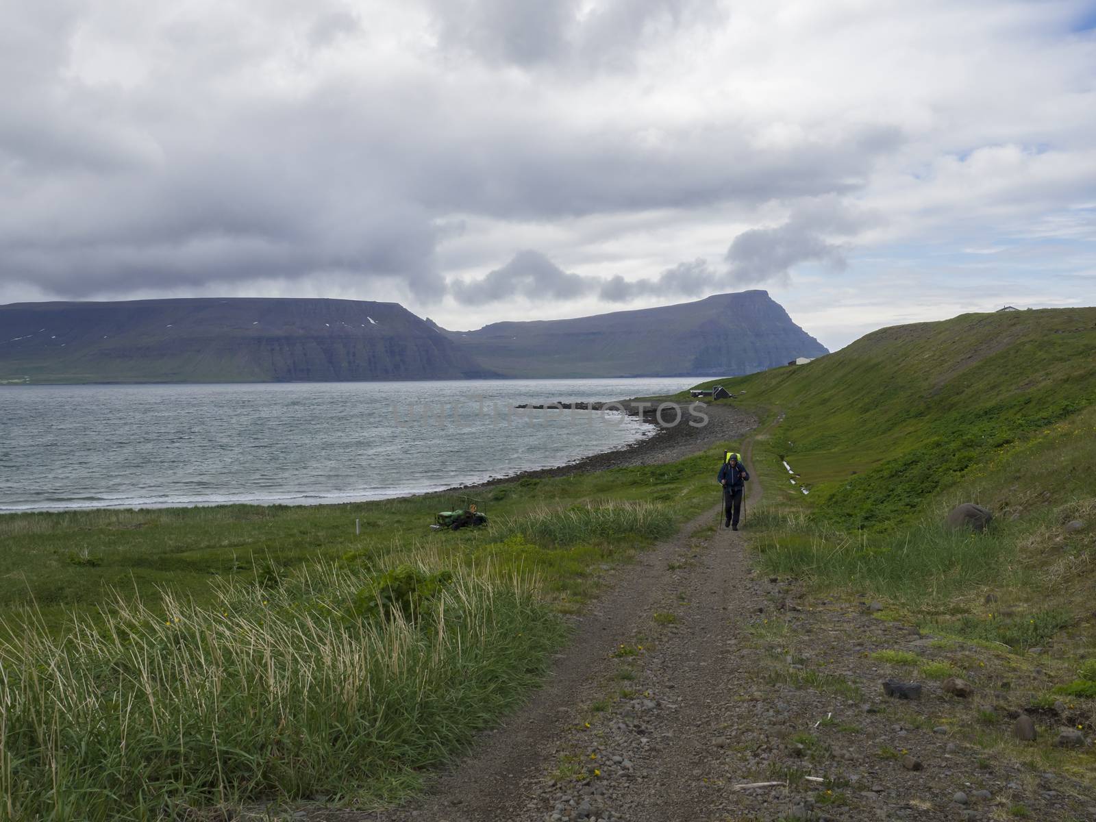 Iceland, West fjords, Hornstrandir, Latrar, June 26, 2018: Lonely man hiker with heavy backpack walking on footpath trail in adalvik cove in nature reserve Hornstrandir, with summer cottages, green grass meadow, stone beach, ocean, snow patched cliffs and blue sky clouds background