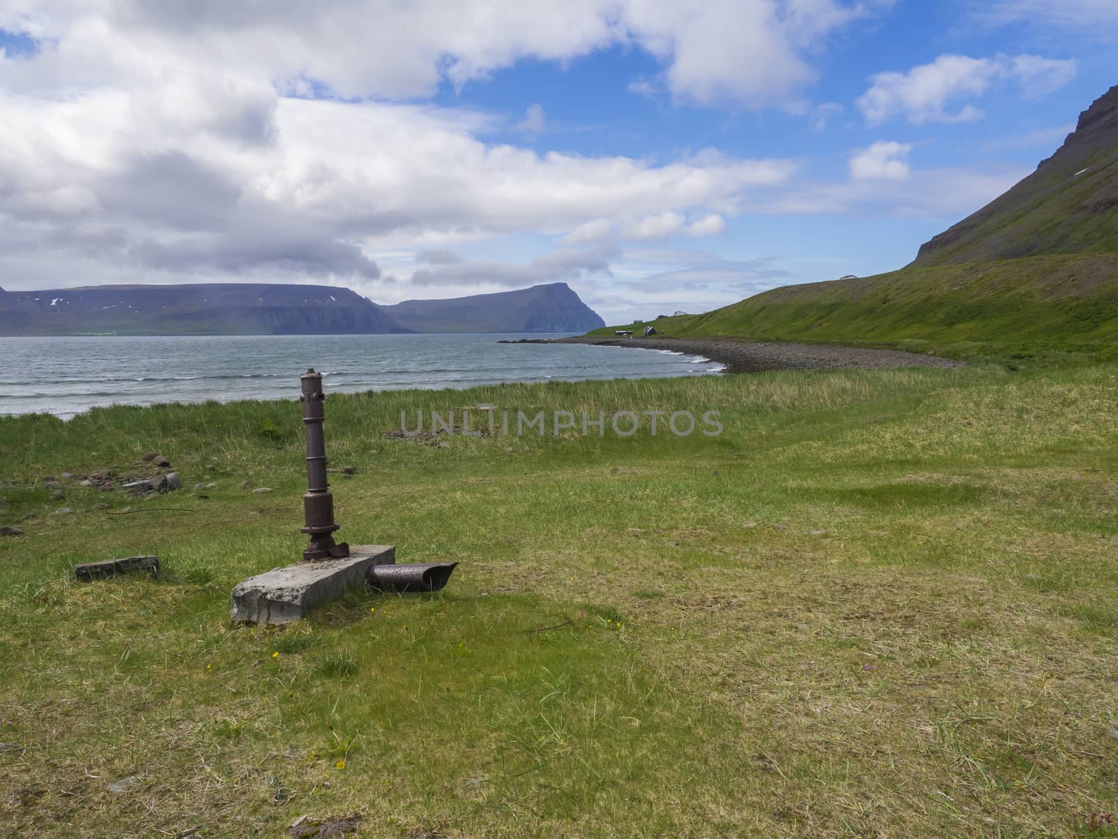View on adalvik cove with rusty water pump and summer cottages at latrar in west fjords nature reserve Hornstrandir in Iceland, with green grass meadow, stone beach, ocean, snow patched hills and blue sky clouds background