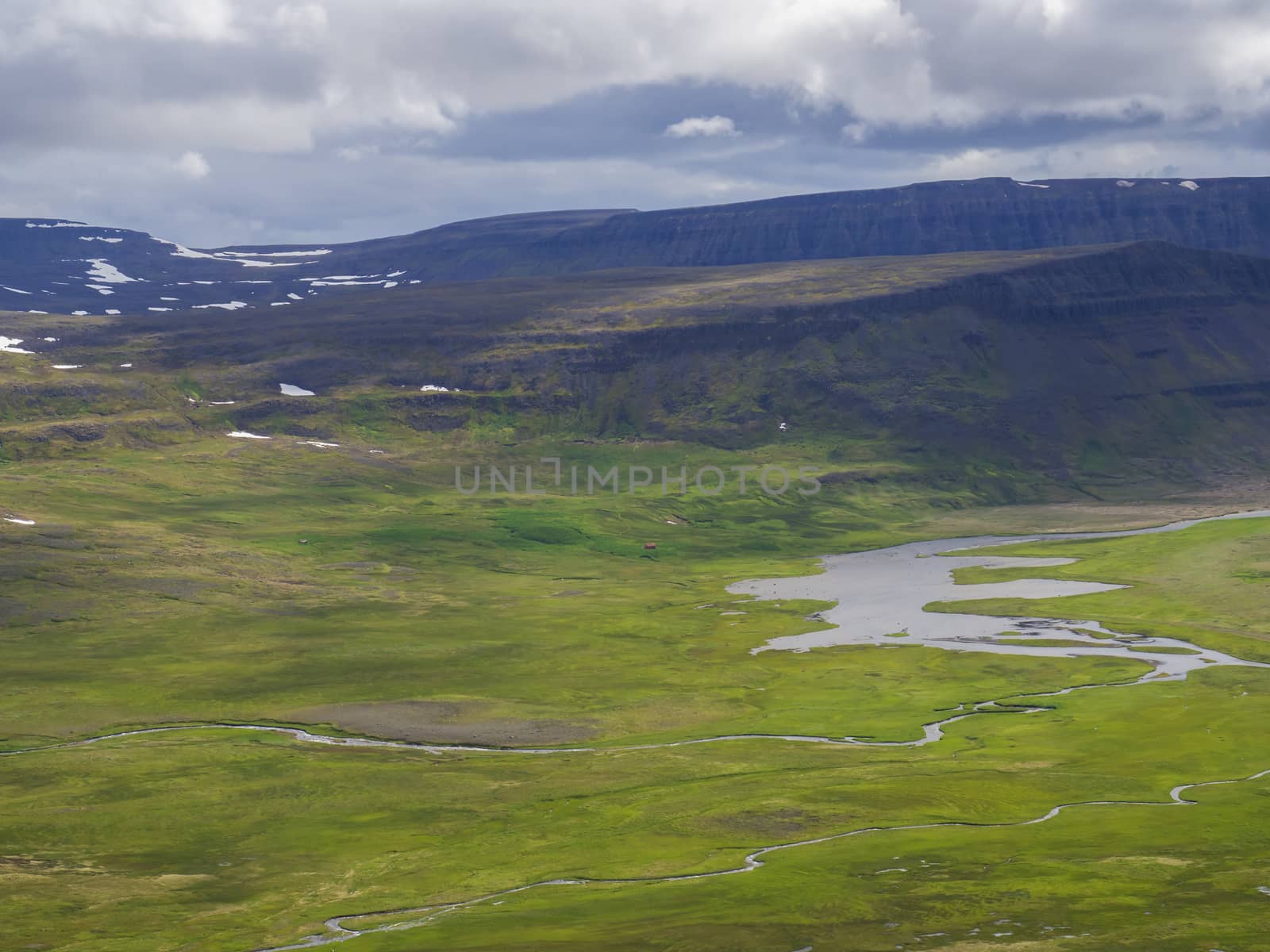 View on beautiful cliffs in adalvik cove in west fjords nature reserve Hornstrandir in Iceland, with river stream delta, red house, snow patched green meadow and dark clouds background by Henkeova