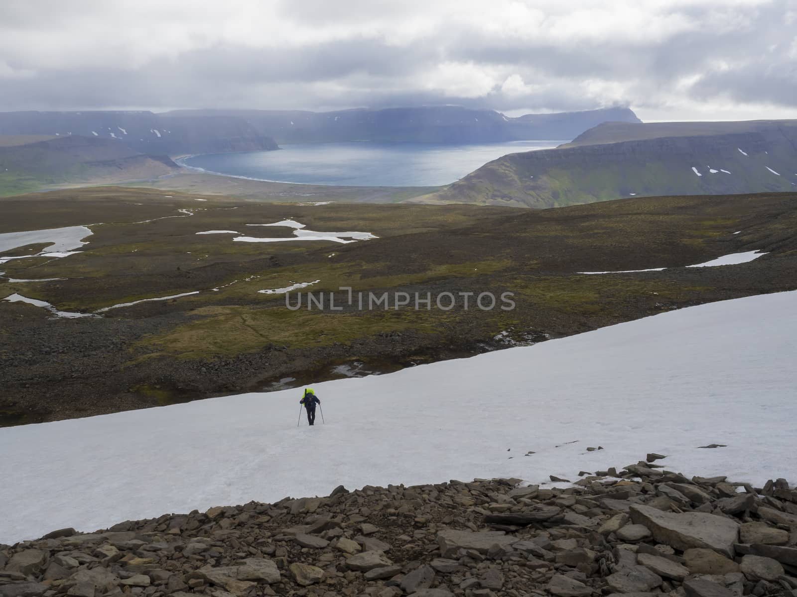 Lonely man hiker with heavy backpack walking on snow field in west fjords nature reserve Hornstrandir in Iceland with view on adalvik cove, ocean, hills and dramatic cliffs, dark cloudy sky background