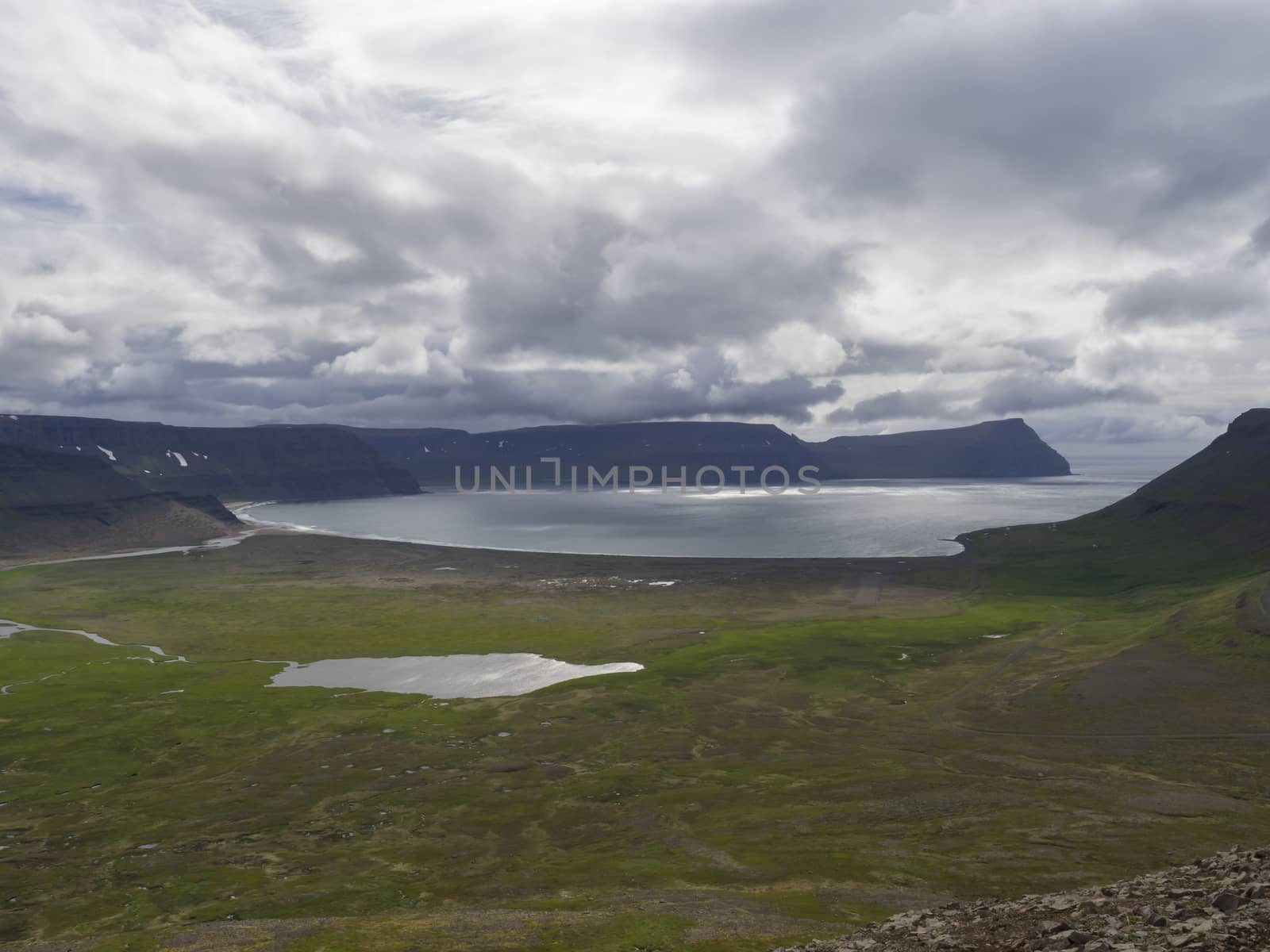 View on adalvik and latrar in west fjords nature reserve Hornstrandir in Iceland with lake and river stream, green grass meadow, beach, ocean, hills and dramatic cliffs, dark cloudy sky background by Henkeova