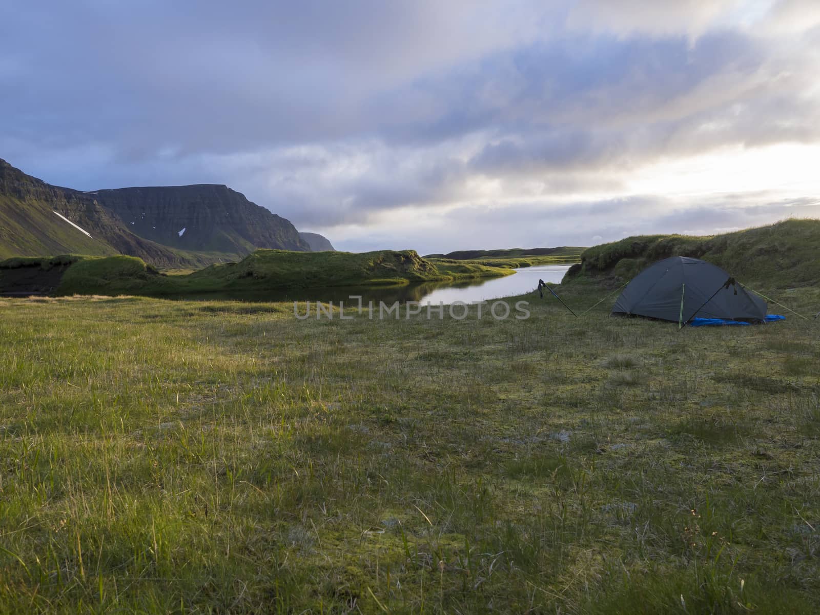 small blue tent standing alone on green grass on mossed creek banks in Hornstrandir Iceland, snow patched hills and cliffs, cloudy sky background, golden hour light, copy space.