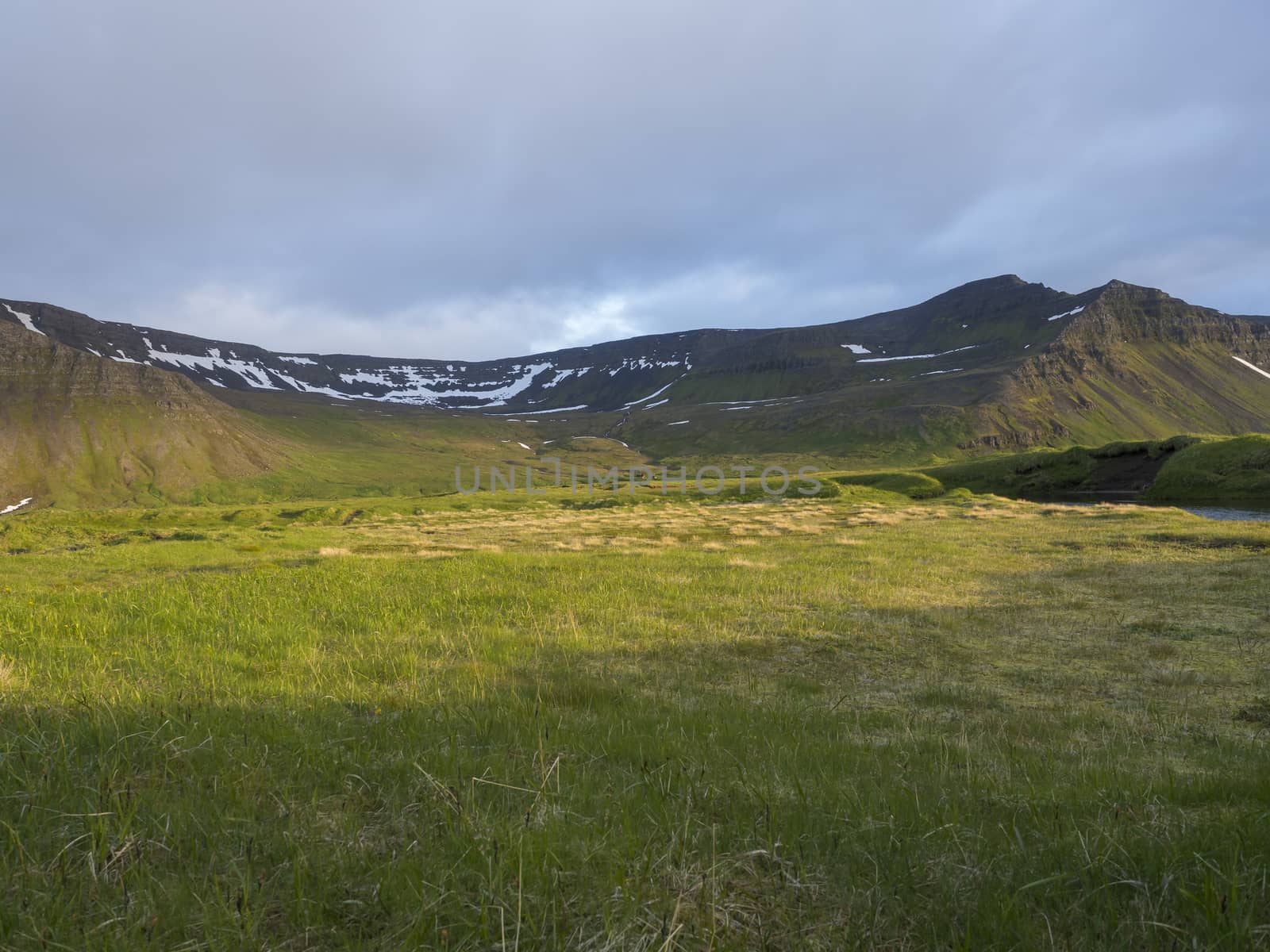 View on beautiful snow covered cliffs mountain and hills in Fljotavik cove, Hornstrandir, west fjords, Iceland, with green grass meadow, golden hour light, blue sky background