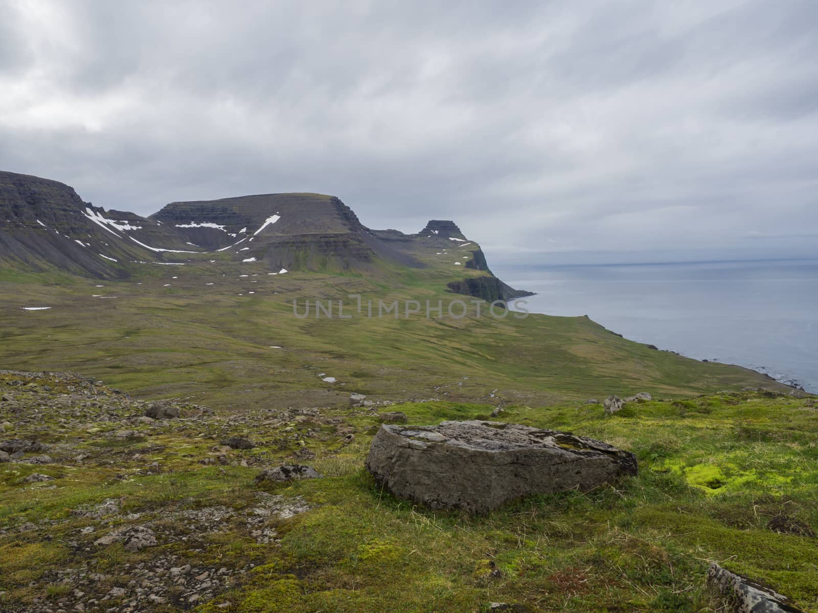 beautiful snow spotted green cliffs and blue sea horizon with big rock stone and green grass meadow, moody sky background, Hornstrandir, west fjords, Iceland