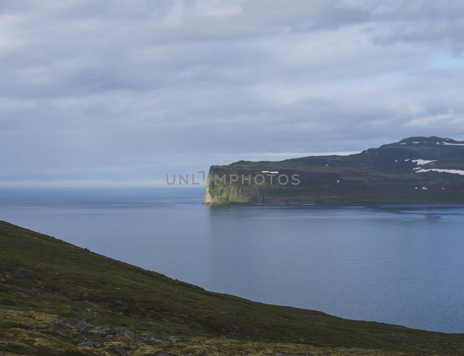Scenic view from ocean on beautiful snow patched cliffs in west fjords, nature reserve Hornstrandir in Iceland, blue sea and cloudy sky background, golden hour light