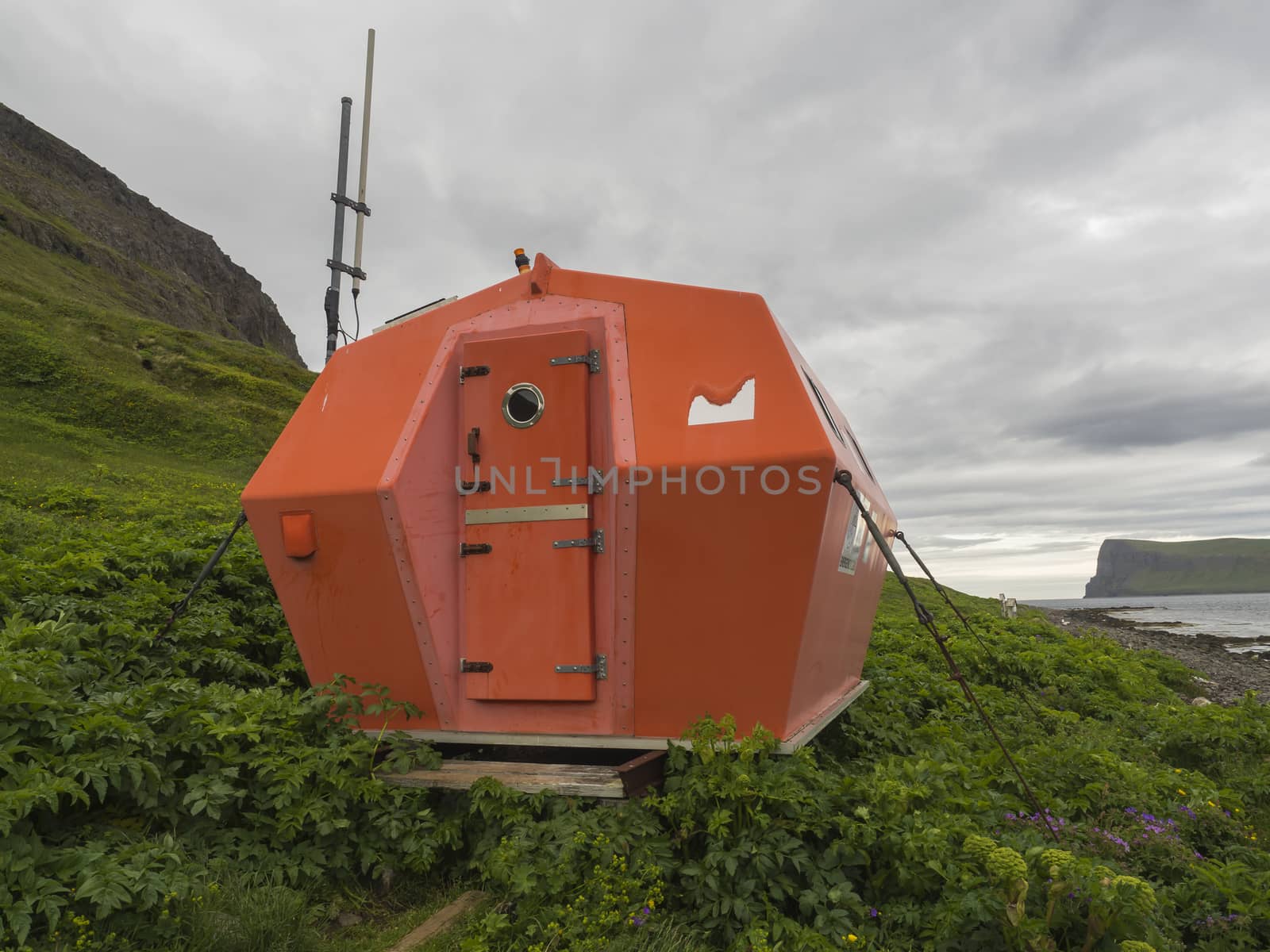 Red emergency shelter cabin in Hornvik standing on the sea shore grass meadow with view on hornbjarg cliffs, Hornstrandir, west fjords, Iceland by Henkeova