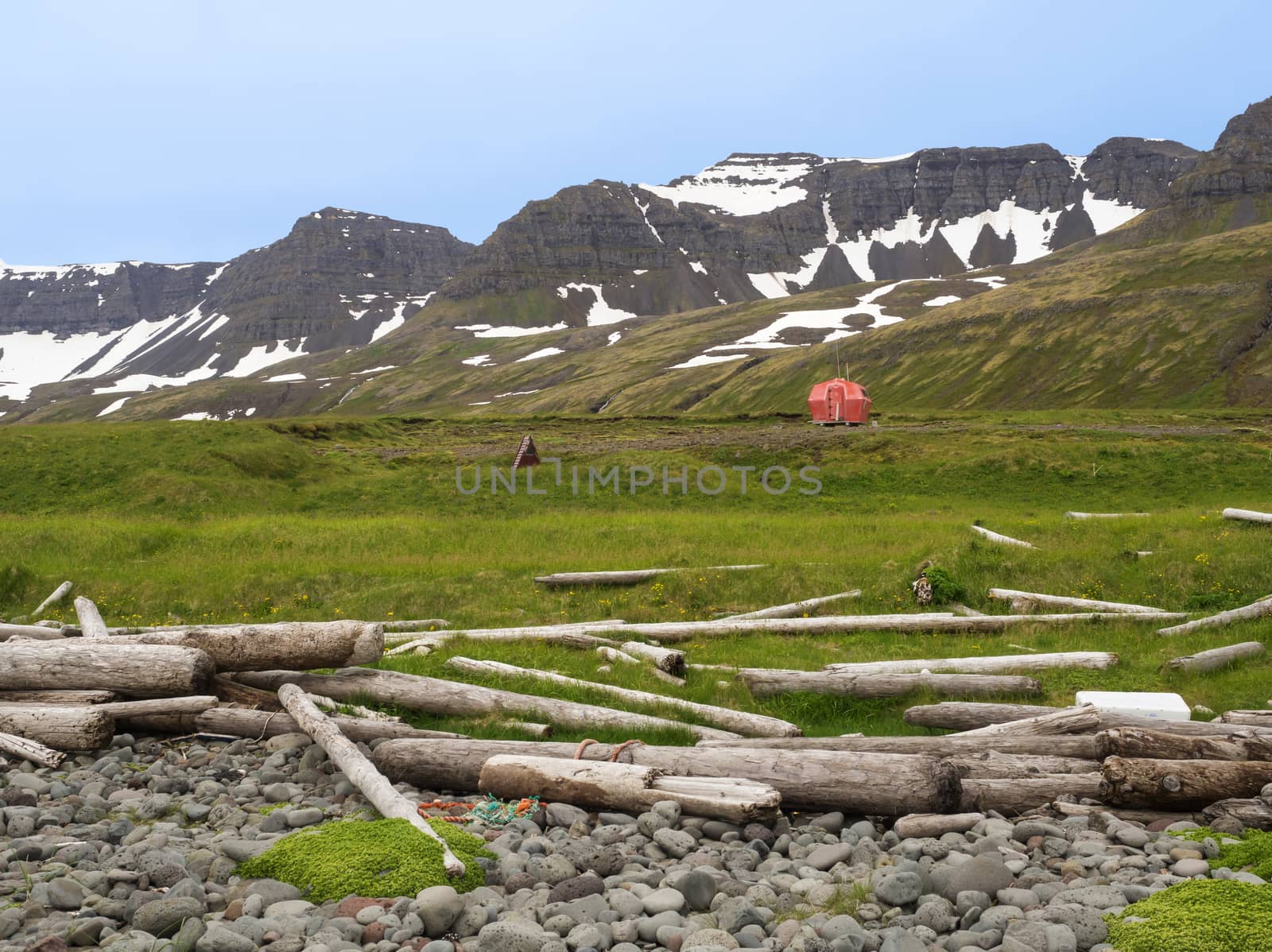Hloduvik campsite with Red emergency shelter cabin standing on the grass meadow with view on snow coved mountain cliffs, stone and wodd logs, Hornstrandir, west fjords, Iceland