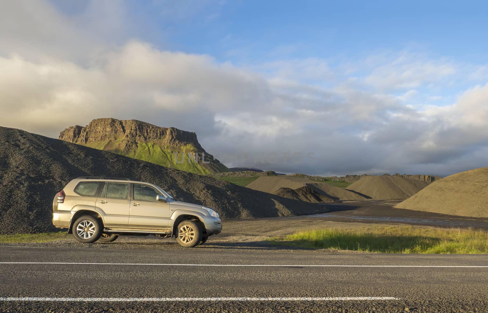 dirty off road car standing next asphalt road with Iceland northern summer landscape, green eroded mountains and blue sky, white clouds by Henkeova