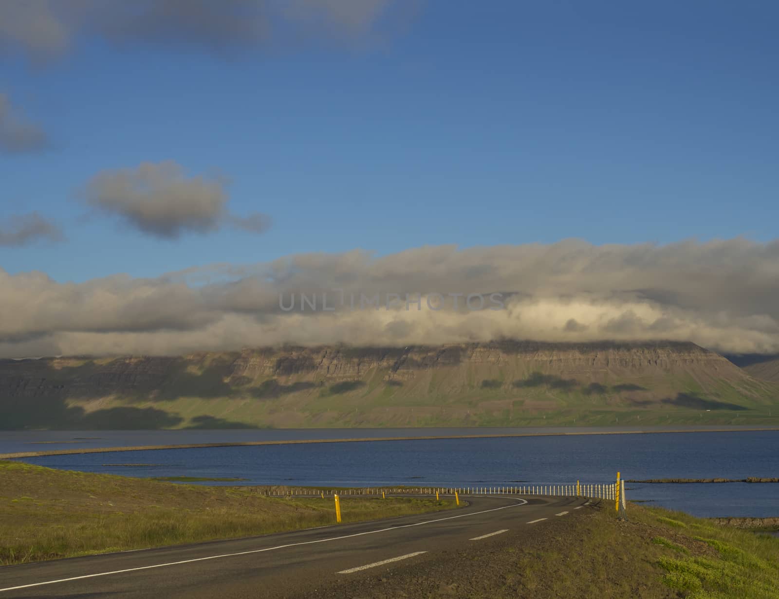 asphalt road through rural northern landscape with green grass and volcanic hills, sea, blue sky clouds, golden hour, summer in Iceland west fjords by Henkeova