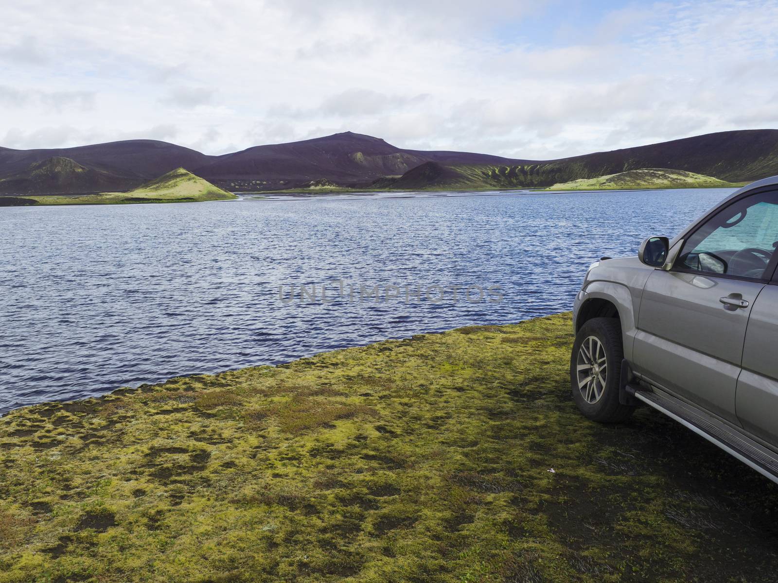 4x4 off road car standing on banks of blue crater lake and volcanic purple and green mountains in Veidivotn lakes, popular fishing area for local, central Iceland highlands in the middle of black lava desert.