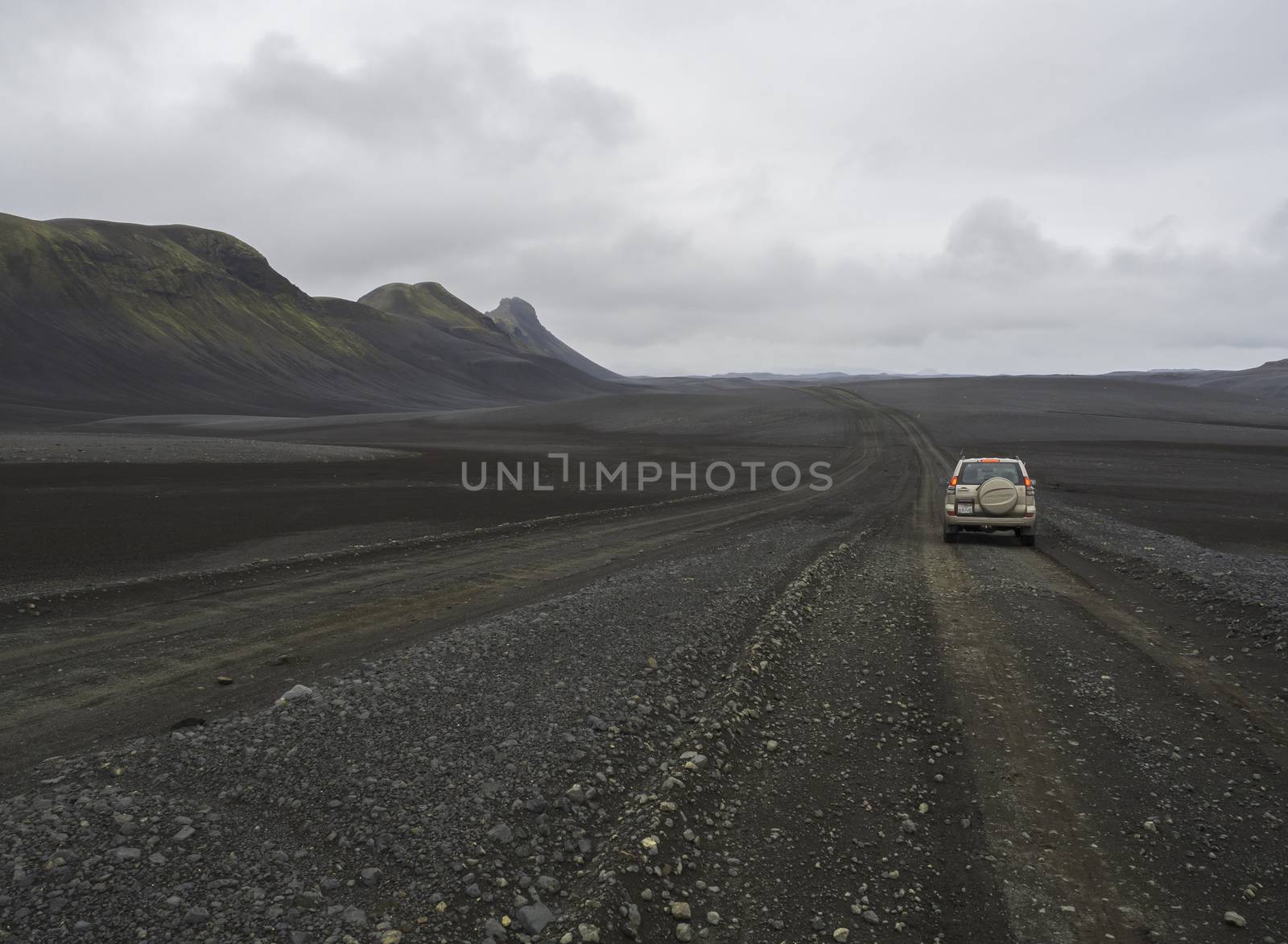 South Iceland, Nature reserve Fjallabak, July 4, 2018: Off road car Toyota Landcruiser driving on dirty mountain road through black lava sand desert landscape with green hills and moody sky by Henkeova