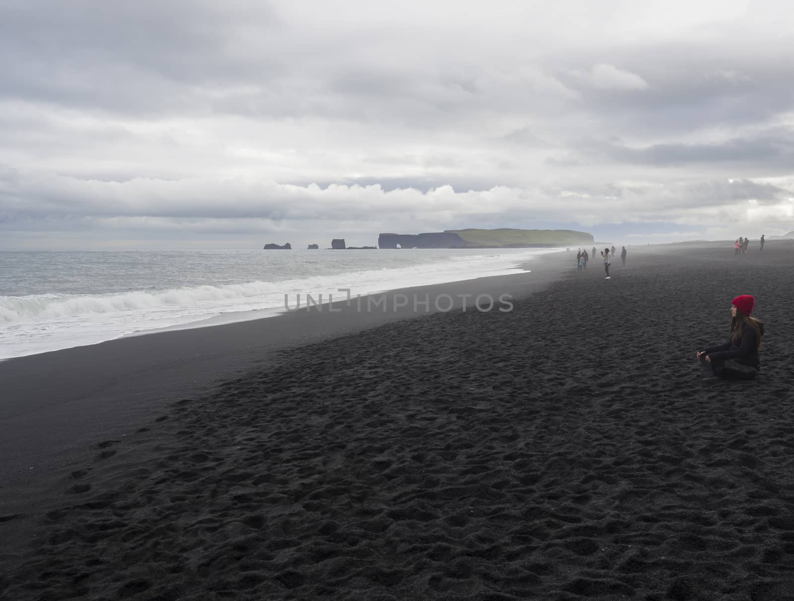 South Iceland, Vik i Myrdal, July 4, 2018: Young pretty woman in red hat sitting meditating or relaxing on Reynisfjara black sand beach, Dyrholaey rock and group of tourist people in backgrund, fog and moody sky, monochromatic look by Henkeova