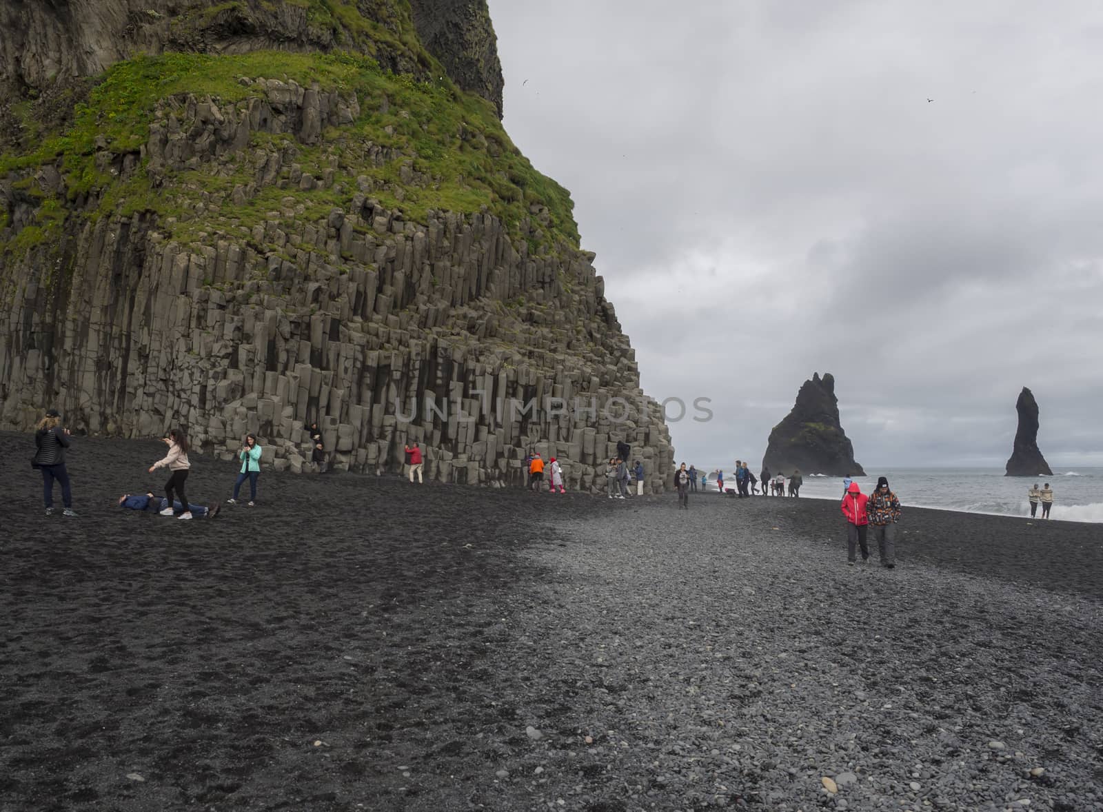 South Iceland, Vik i Myrdal, July 4, 2018: Group of colorful dressed tourist people takeing pictures and havenig fun at Basalt rock columns and Troll toes on black sand beach reynisfjara, fog and moody sky, monochromatic look