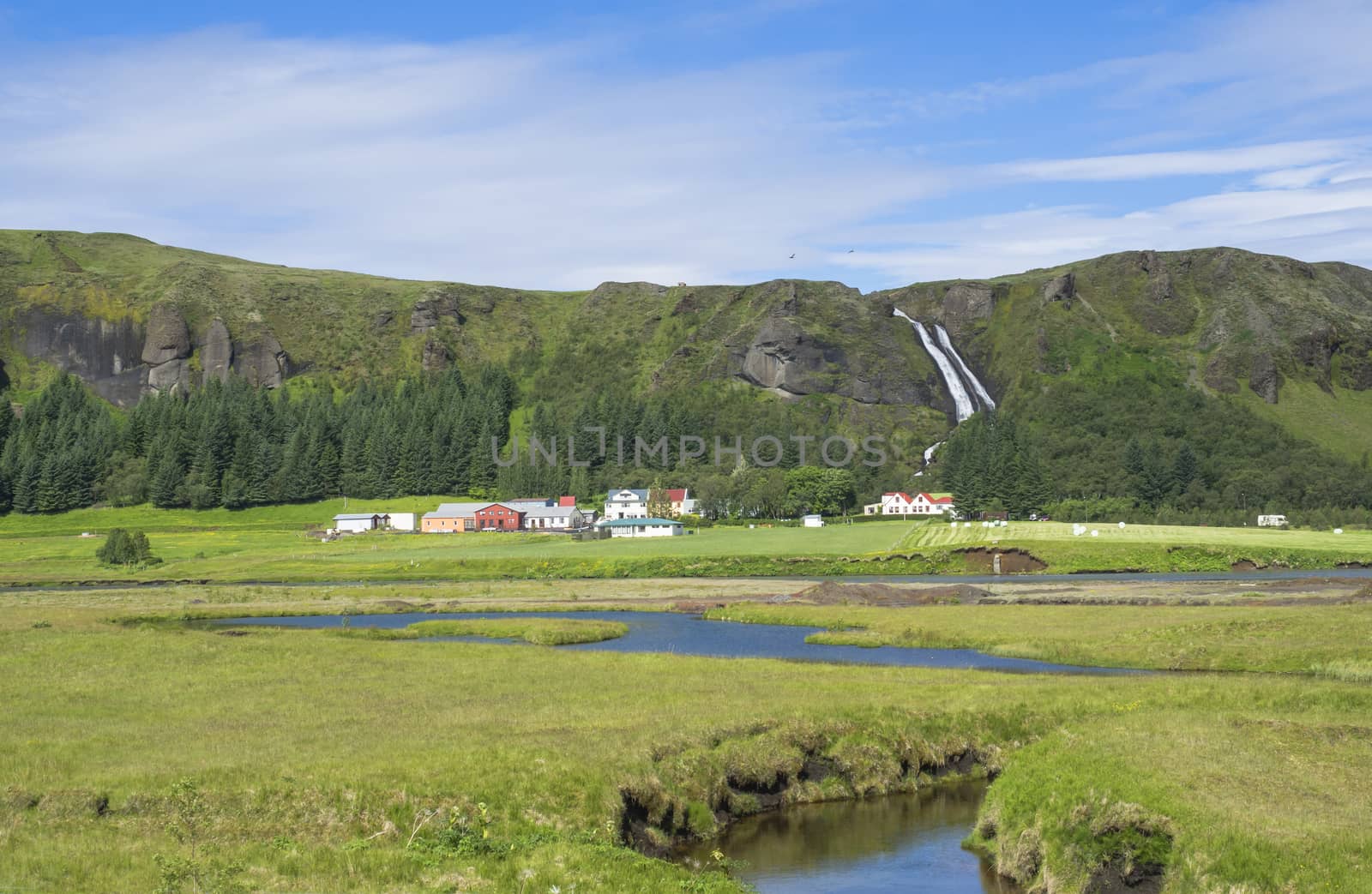 rural landscape with colorful farm houses, blue river stream on green grass meadow with spruce tree forest, hills and beatuful waterfall Systrafoss in Kirkjub�jarklaustur, Iceland