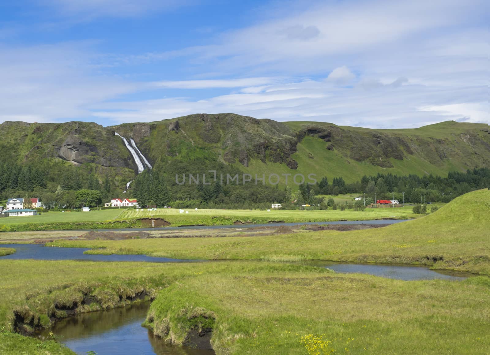 rural landscape with colorful farm houses, blue river stream on green grass meadow with spruce tree forest, hills and beatuful waterfall Systrafoss in Kirkjubeajarklaustur, Iceland