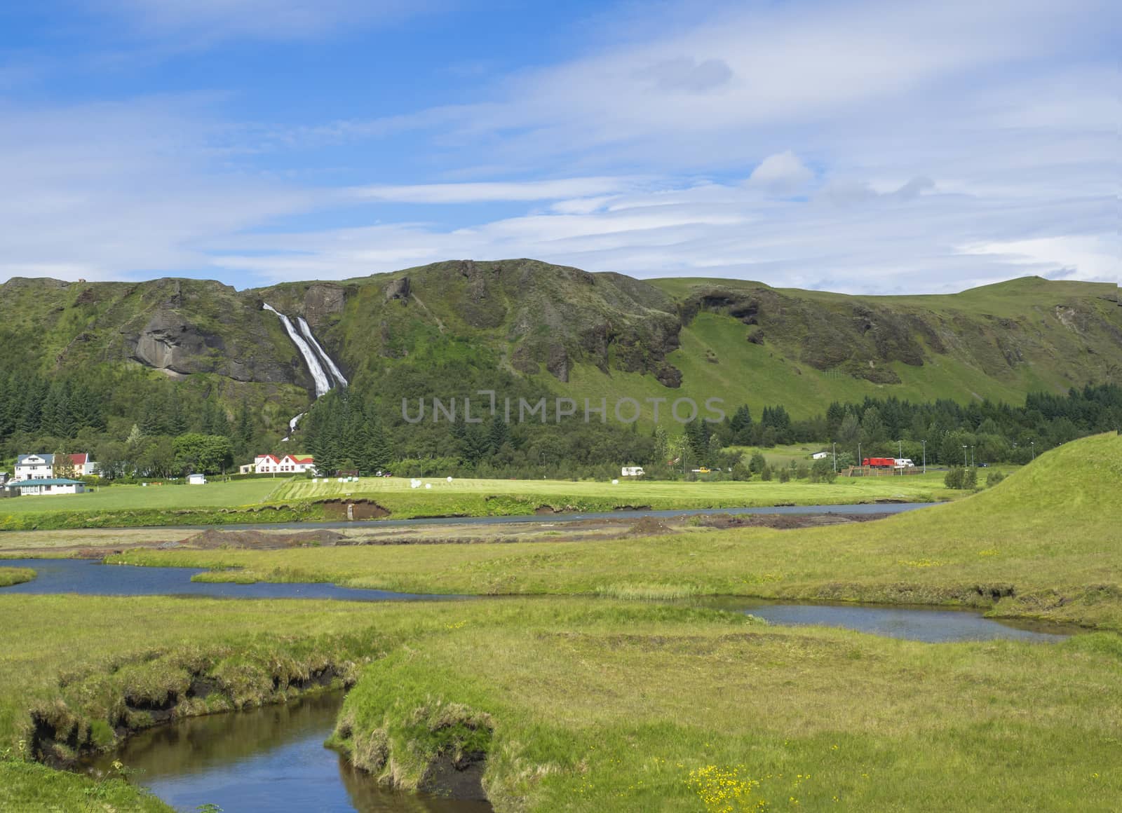 rural landscape with colorful farm houses, blue river stream on green grass meadow with spruce tree forest, hills and beatuful waterfall Systrafoss in Kirkjubeajarklaustur by Henkeova