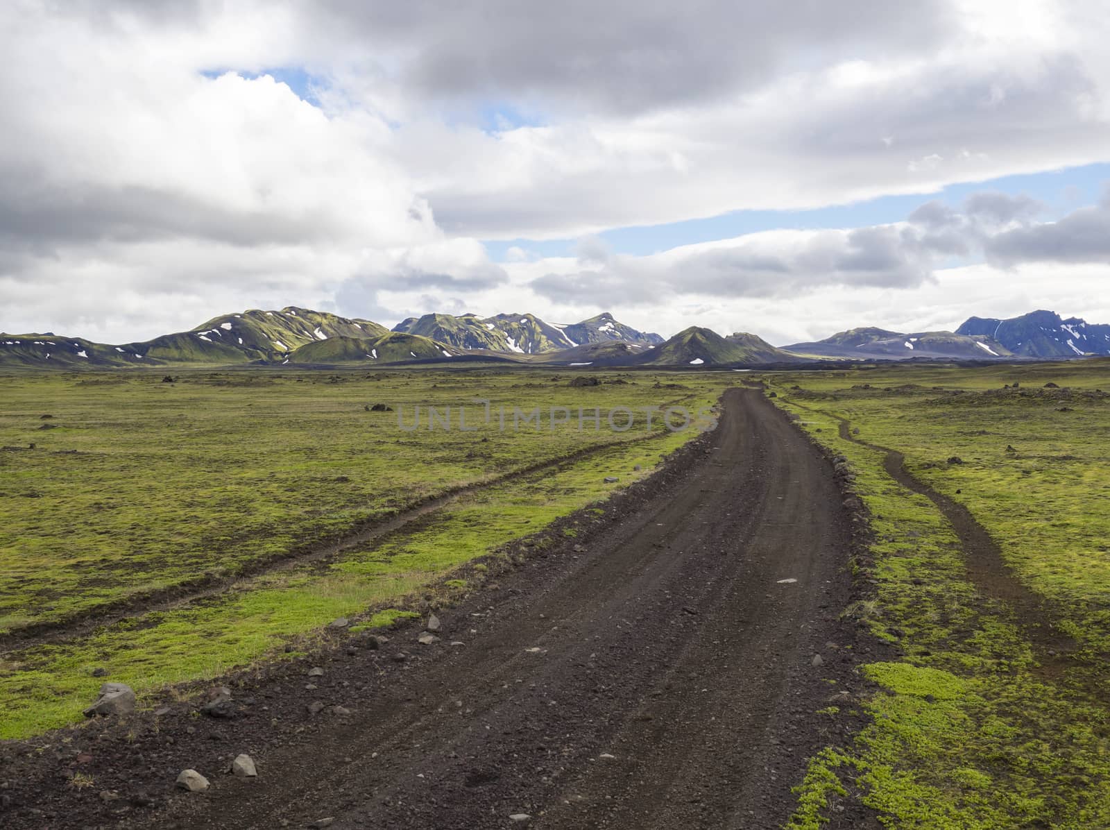 view on dirt mountain road F210 in abandoned green landscape at Nature reserve Fjallabak in Iceland with colorful snow covered rhyolit mountains, blue sky white clouds