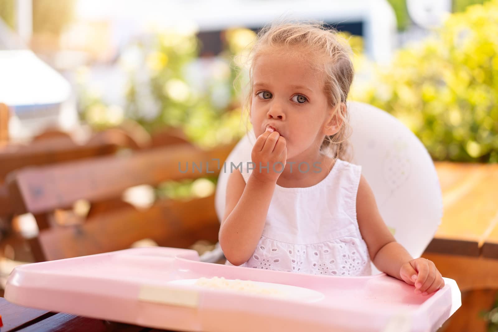 Cute little Caucasian girl eating spaghetti at table sitting in child seat outdoor restaurant. Healthy eating. Funny child eat street food sunny summer day.