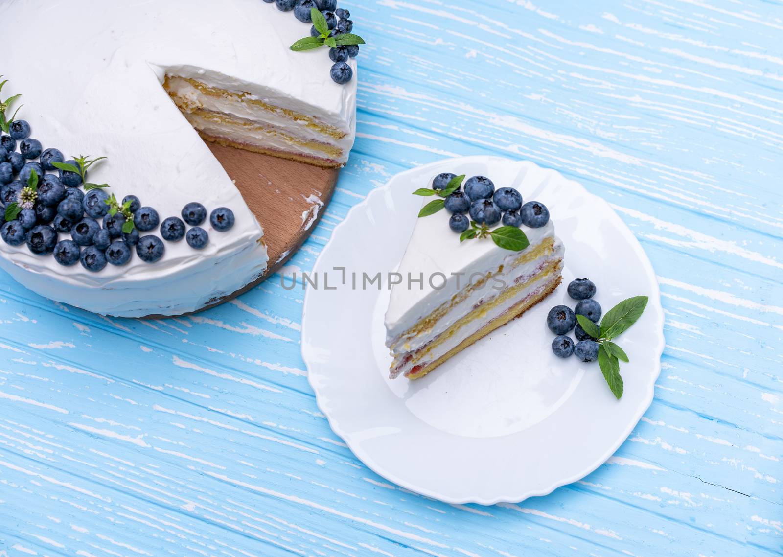 Appetizing cheesecake biscuit pillow decorated white cream blueberries and mint stands on wooden blue rustic table. Sweet cake with piece and spoon on plate