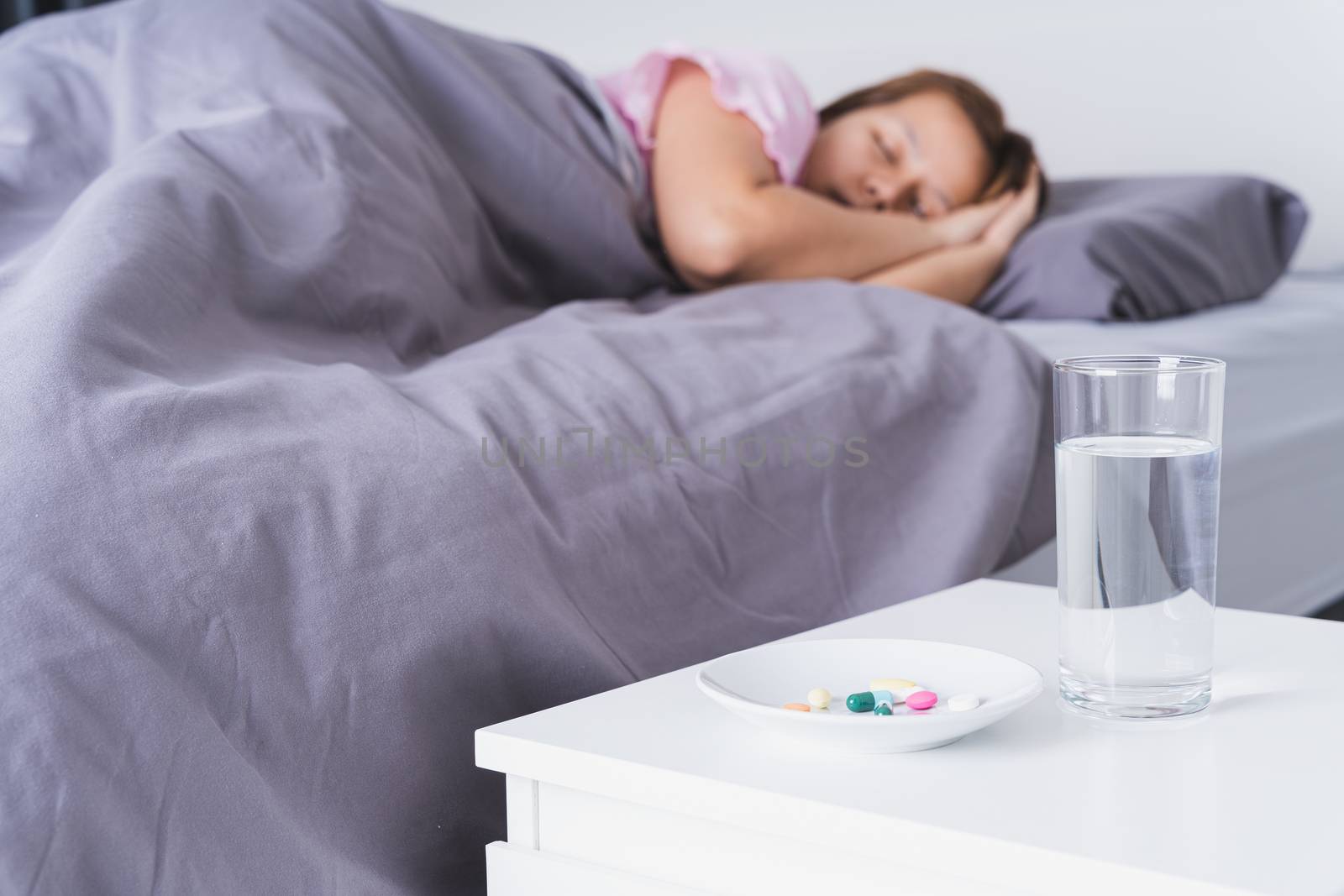 Close up pill drugs in the dish and a glass of water on nightstand and female patient lying on the bed in the bedroom. Healthcare and medical pharmacy concept by mikesaran