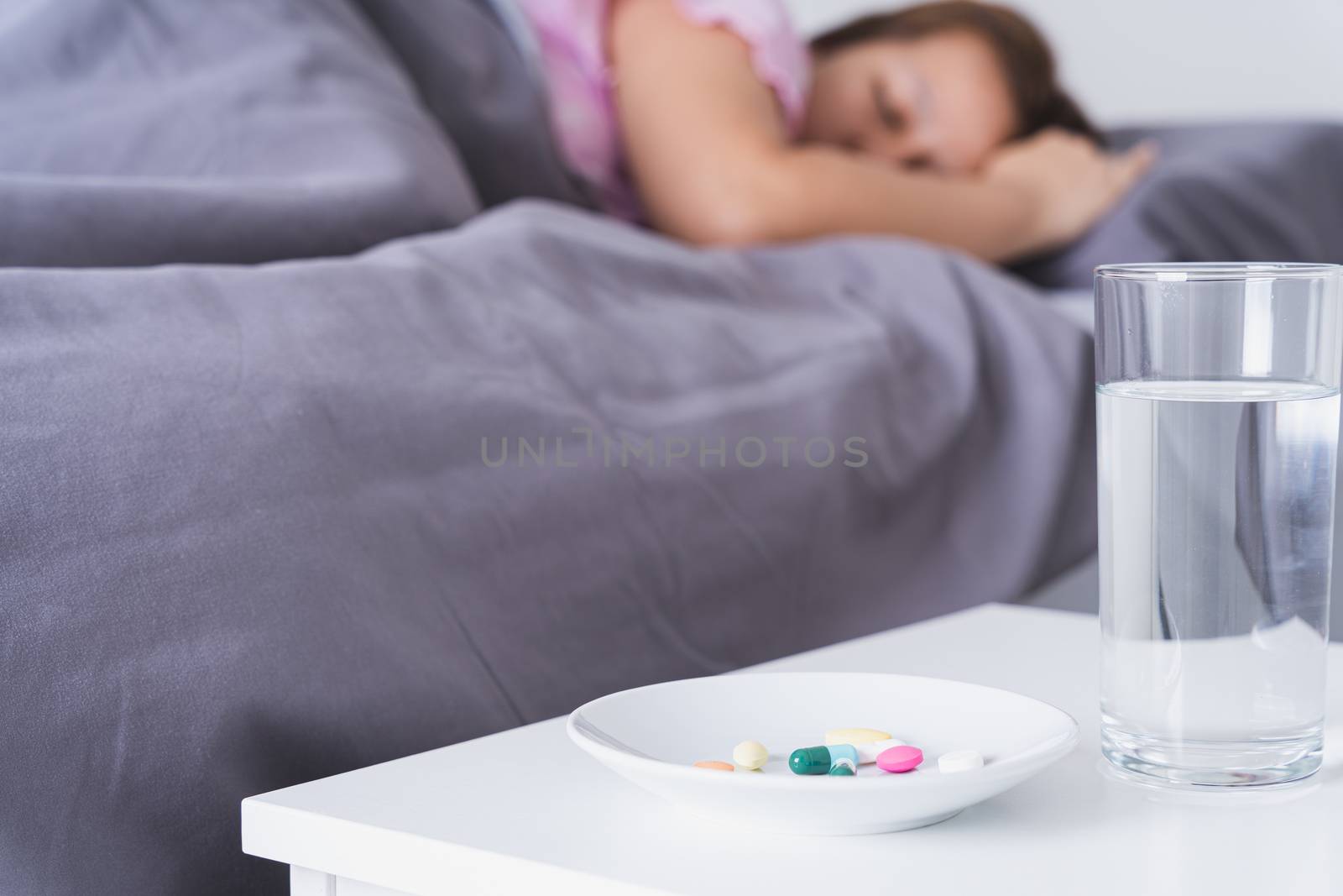 Close up pill drugs in the dish and a glass of water on nightstand and female patient lying on the bed in the bedroom. Healthcare and medical pharmacy concept