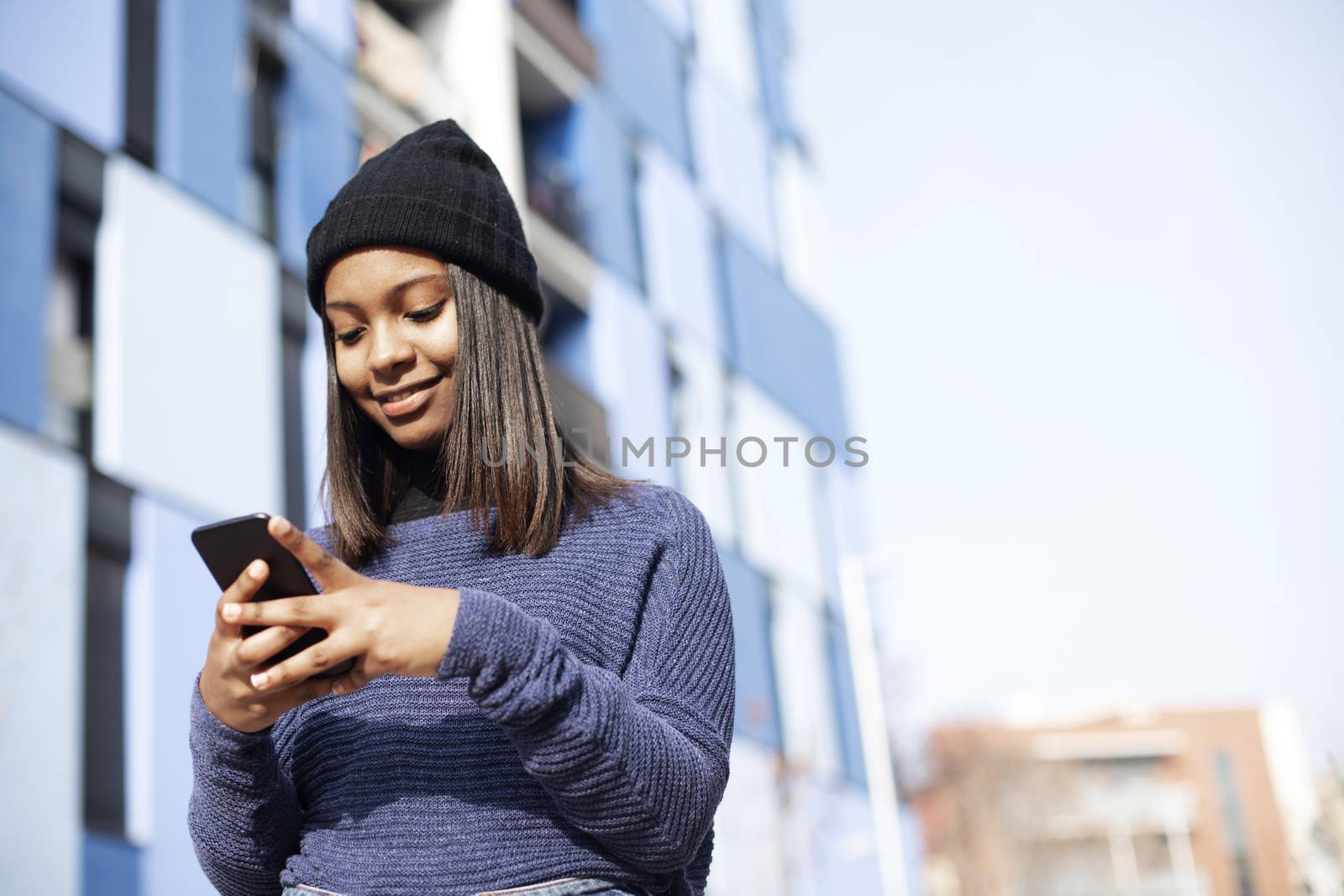 Portrait of African American young beautiful woman wearing a wool cap standing on the street while using a mobile phone by raferto1973