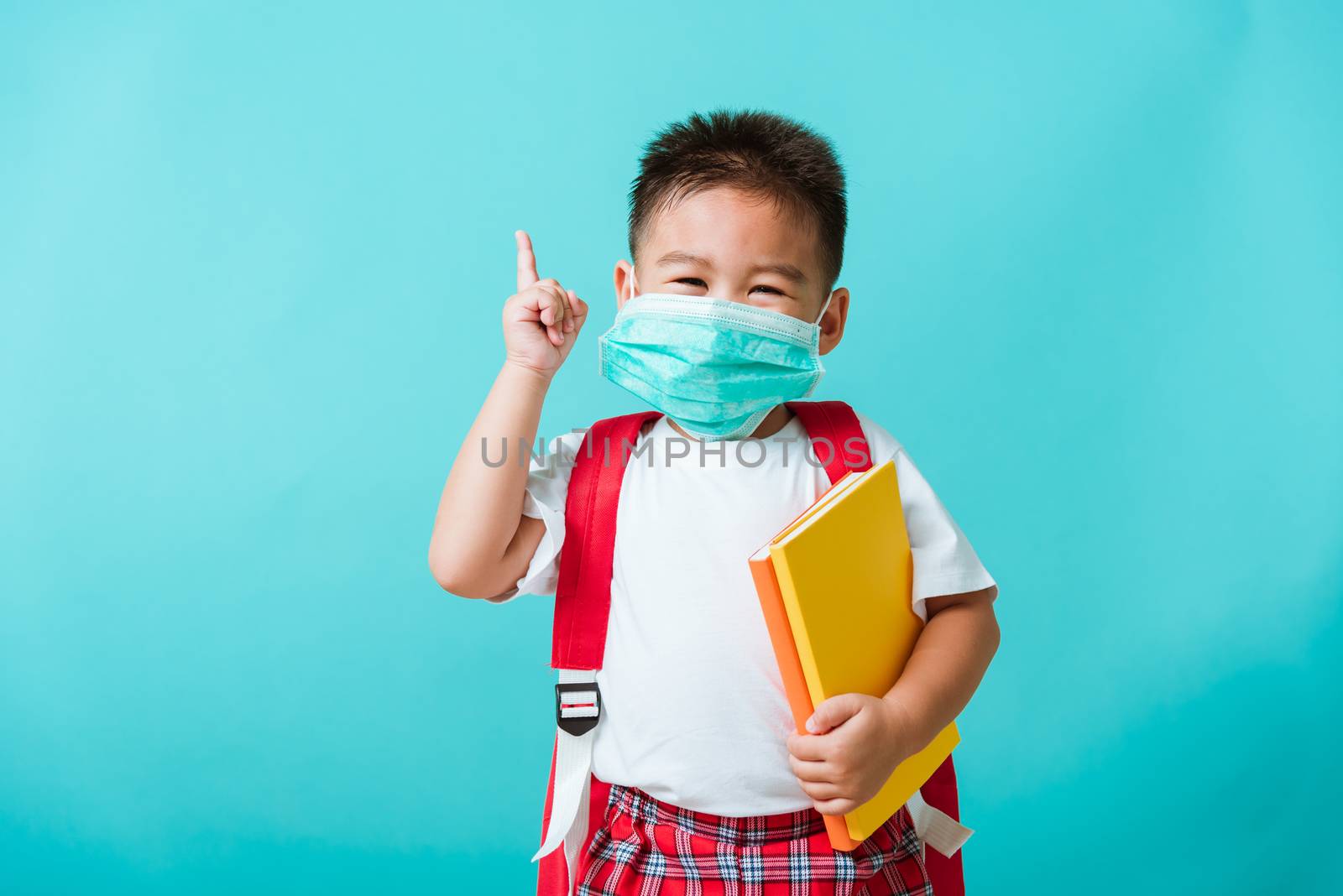 Back to school coronavirus Covid-19 education. Portrait Asian little child boy kindergarten wear face mask protective and school bag hold book before going to school pointing to side away, studio shot