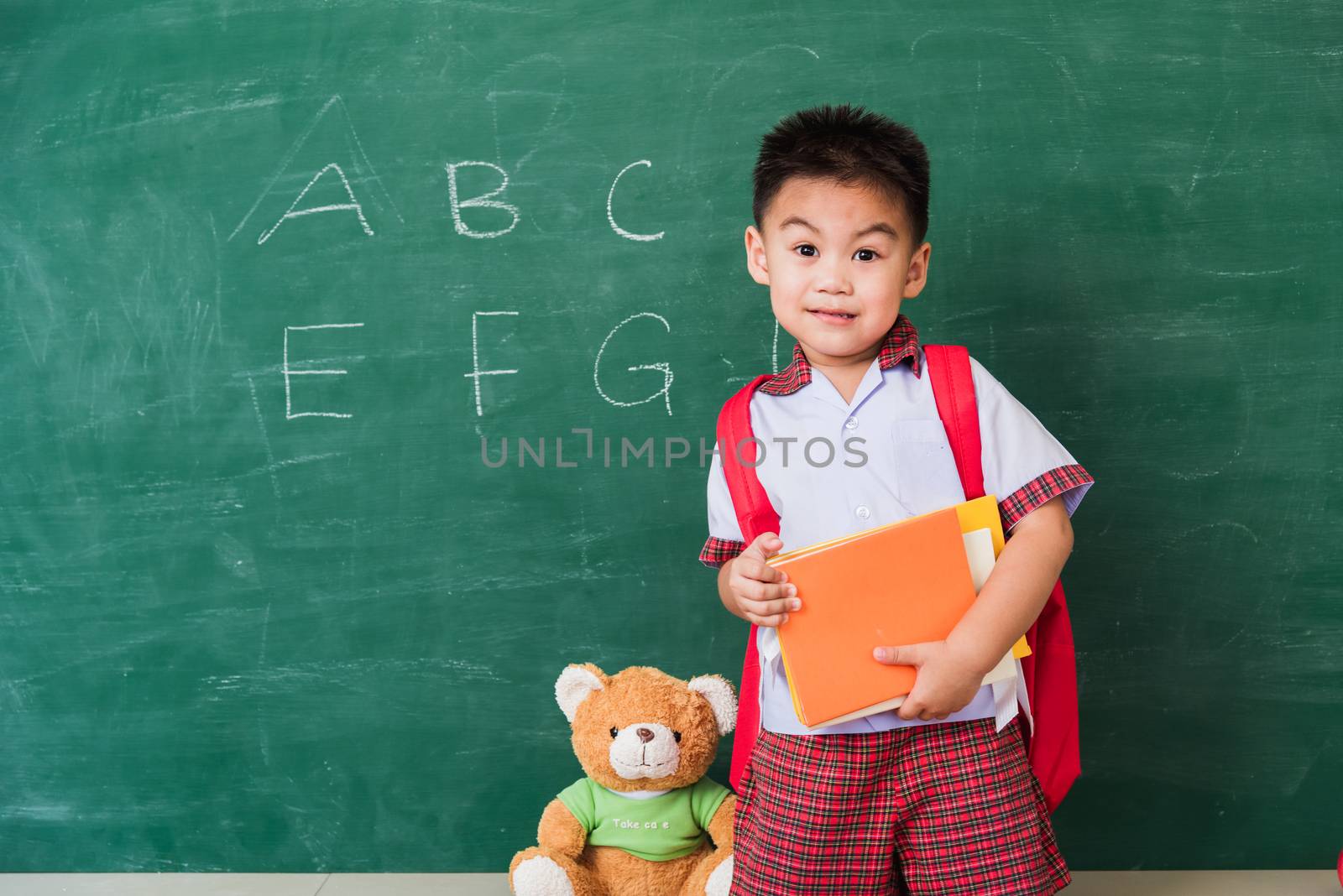 Back to School. Happy Asian funny cute little child boy from kindergarten in student uniform with school bag hold books smile on green school blackboard, First time to school education concept