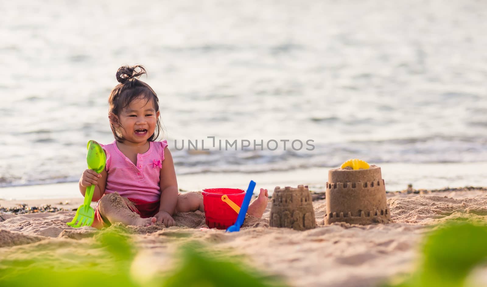 Happy fun Asian child cute little girl playing sand with toy sand tools at a tropical sea beach in holiday summer on sunset time, tourist trip concept