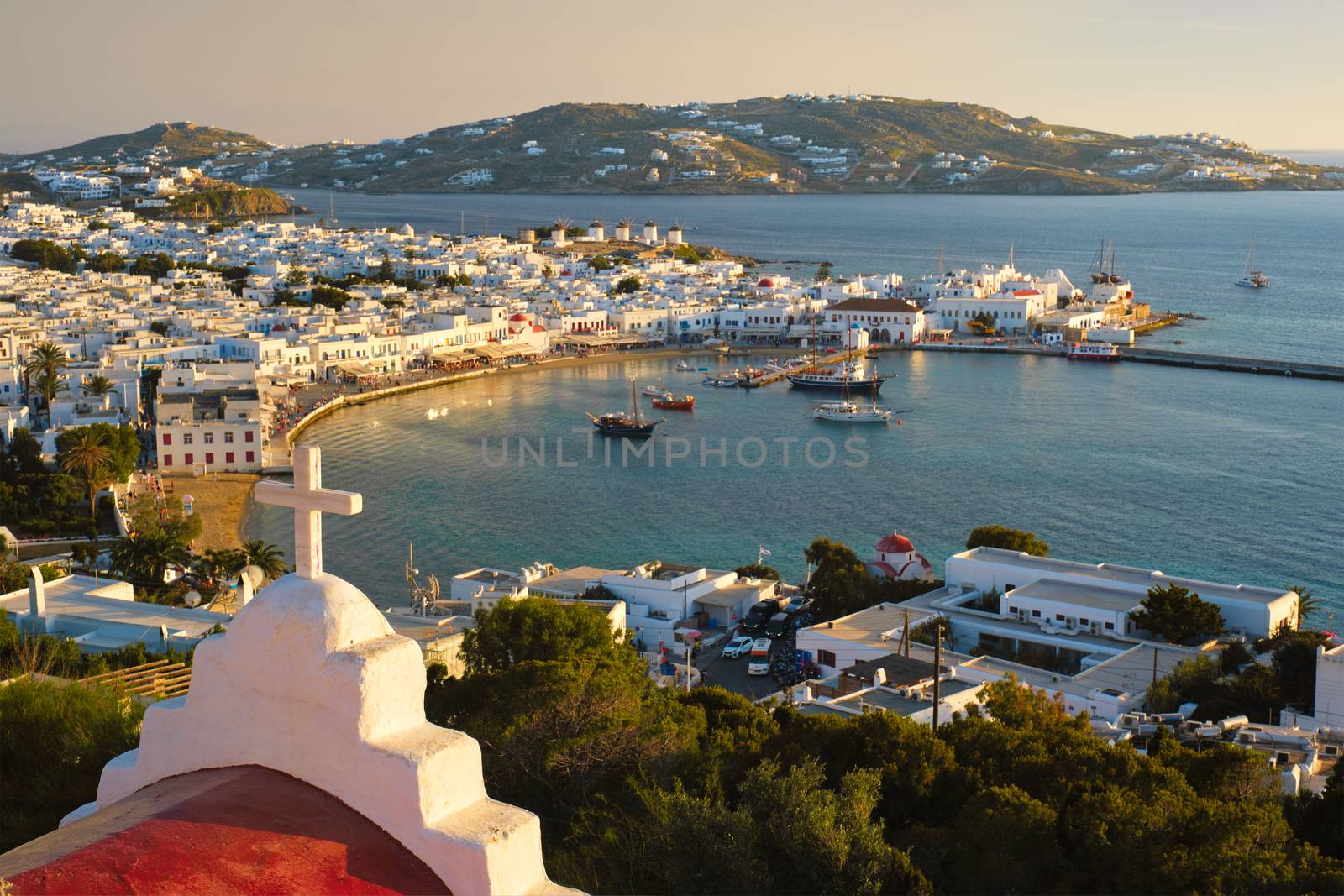 View of Mykonos town Greek tourist holiday vacation destination with famous windmills, and port with boats and yachts on sunset over St Basil church cross. Mykonos, Cyclades islands, Greece.