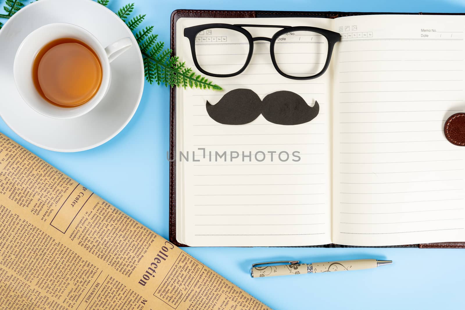 Happy Father Day background concept with notebook, pen, leaf, cup of tea, glasses, black mustache and newspaper on blue background with copy space for text.