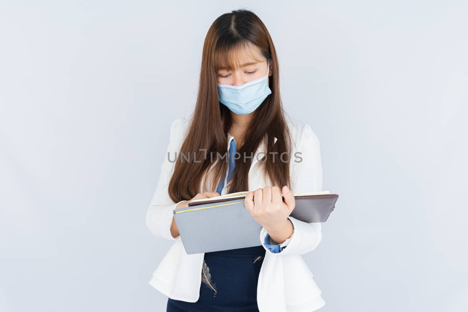 Asian business woman wearing a medical face mask and looking at the notebook that she holding over grey background. Back to the normal concept. by mikesaran