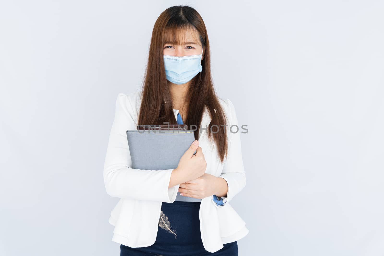 Smiling Asian business woman wearing a medical face mask holding the notebook and looking at the camera over grey background. Back to the normal concept.