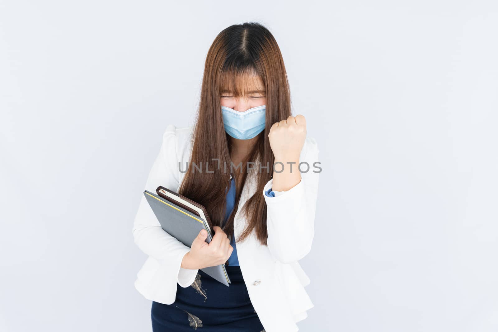 Smiling Asian business woman wearing a medical face mask holding the notebook and showing fist fighting over grey background. Back to the normal concept. by mikesaran