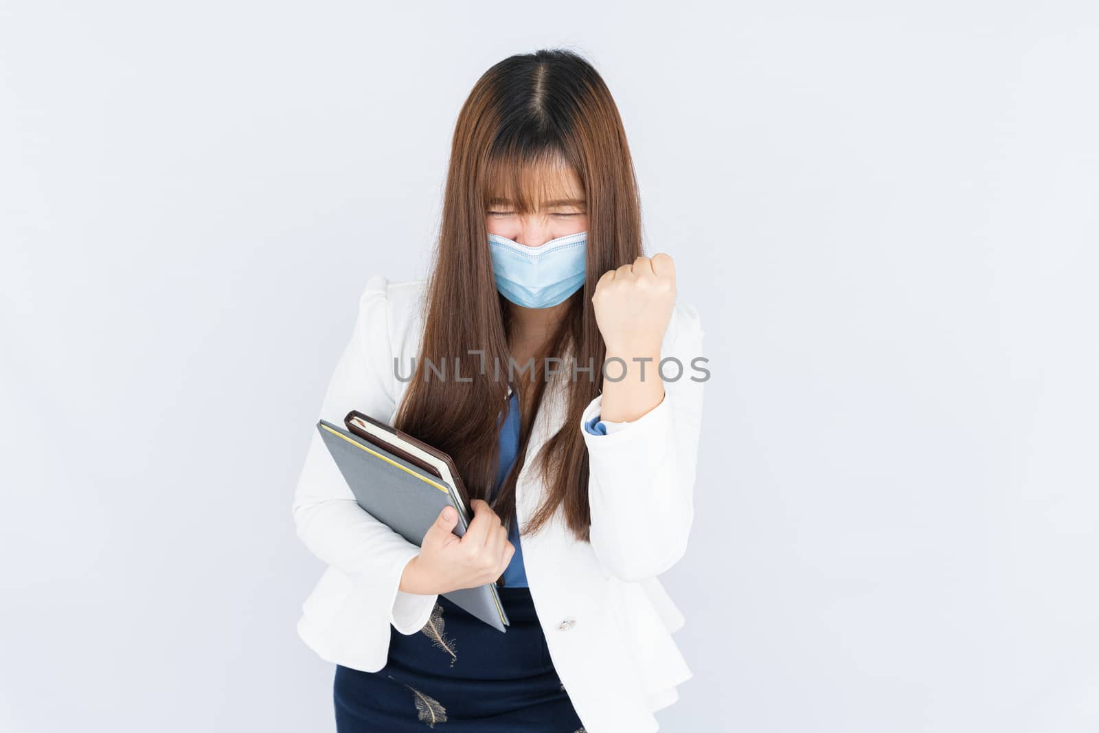 Smiling Asian business woman wearing a medical face mask holding the notebook and showing fist fighting over grey background. Back to the normal concept.