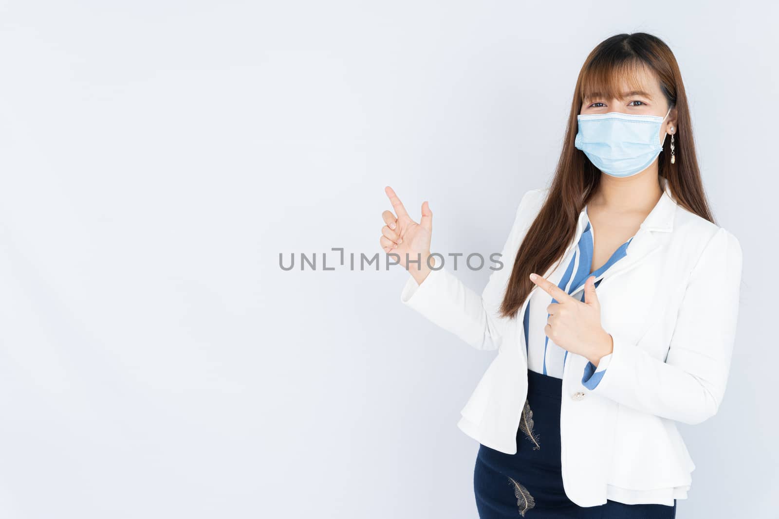 Smiling Asian business woman wearing a medical face mask and pointing finger to side blank space over grey background. Back to the normal concept. by mikesaran