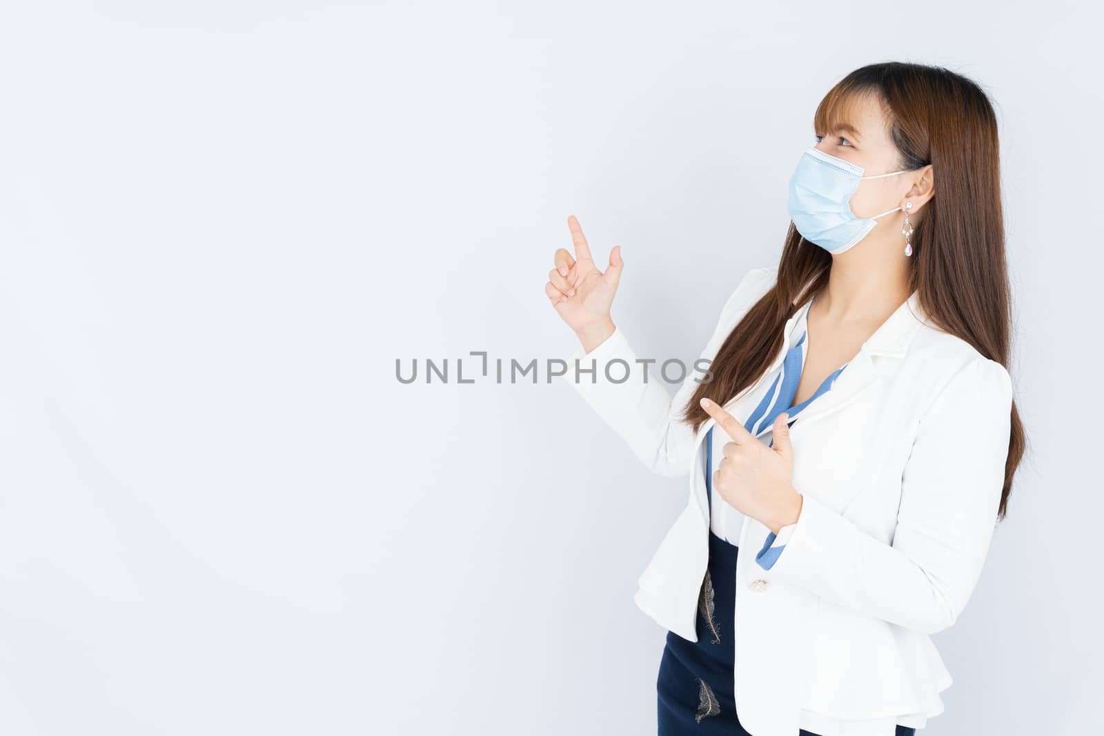Smiling Asian business woman wearing a medical face mask and pointing finger to side blank space over grey background. Back to the normal concept. by mikesaran