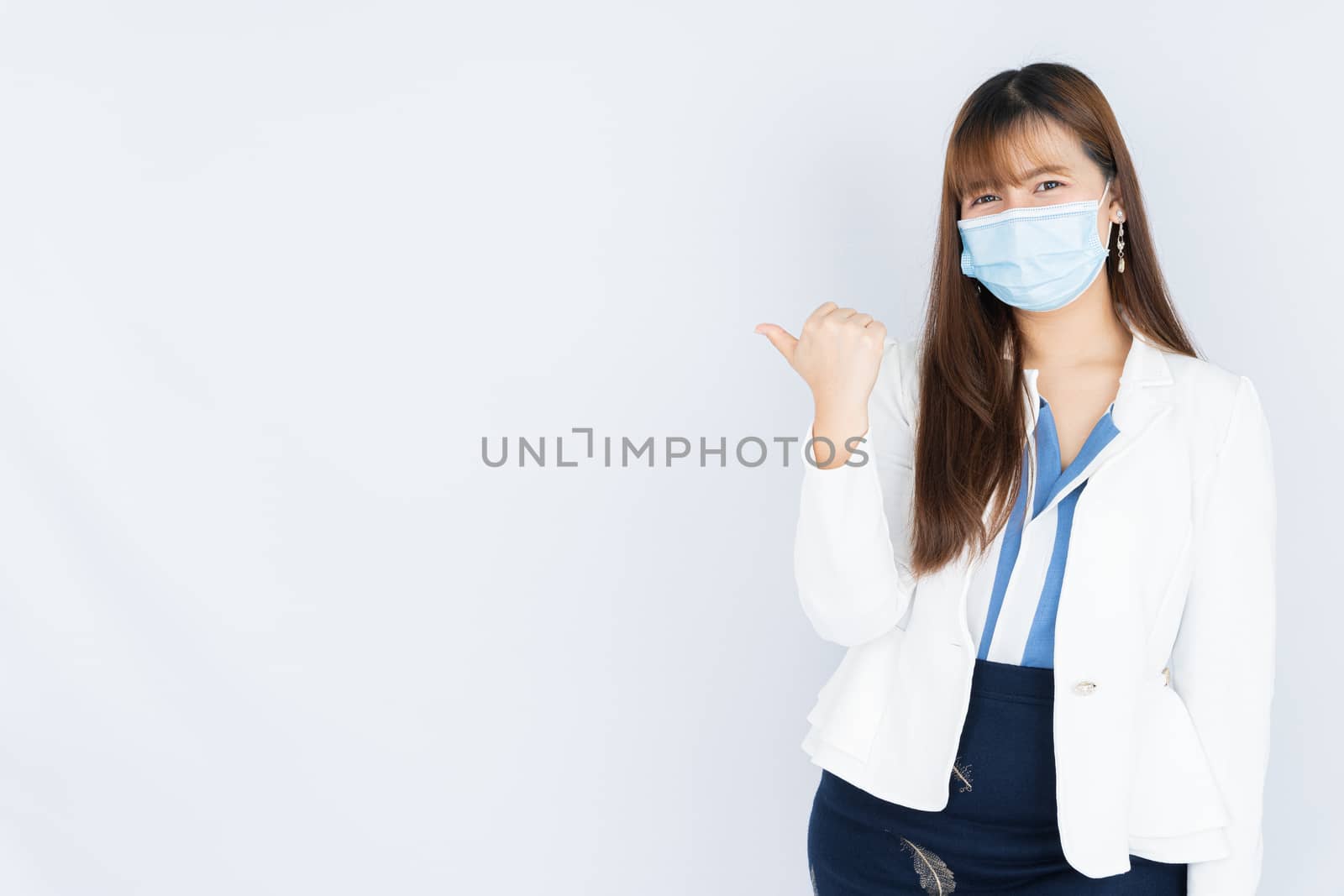 Smiling Asian business woman wearing a medical face mask and pointing finger to side blank space over grey background. Back to the normal concept. by mikesaran