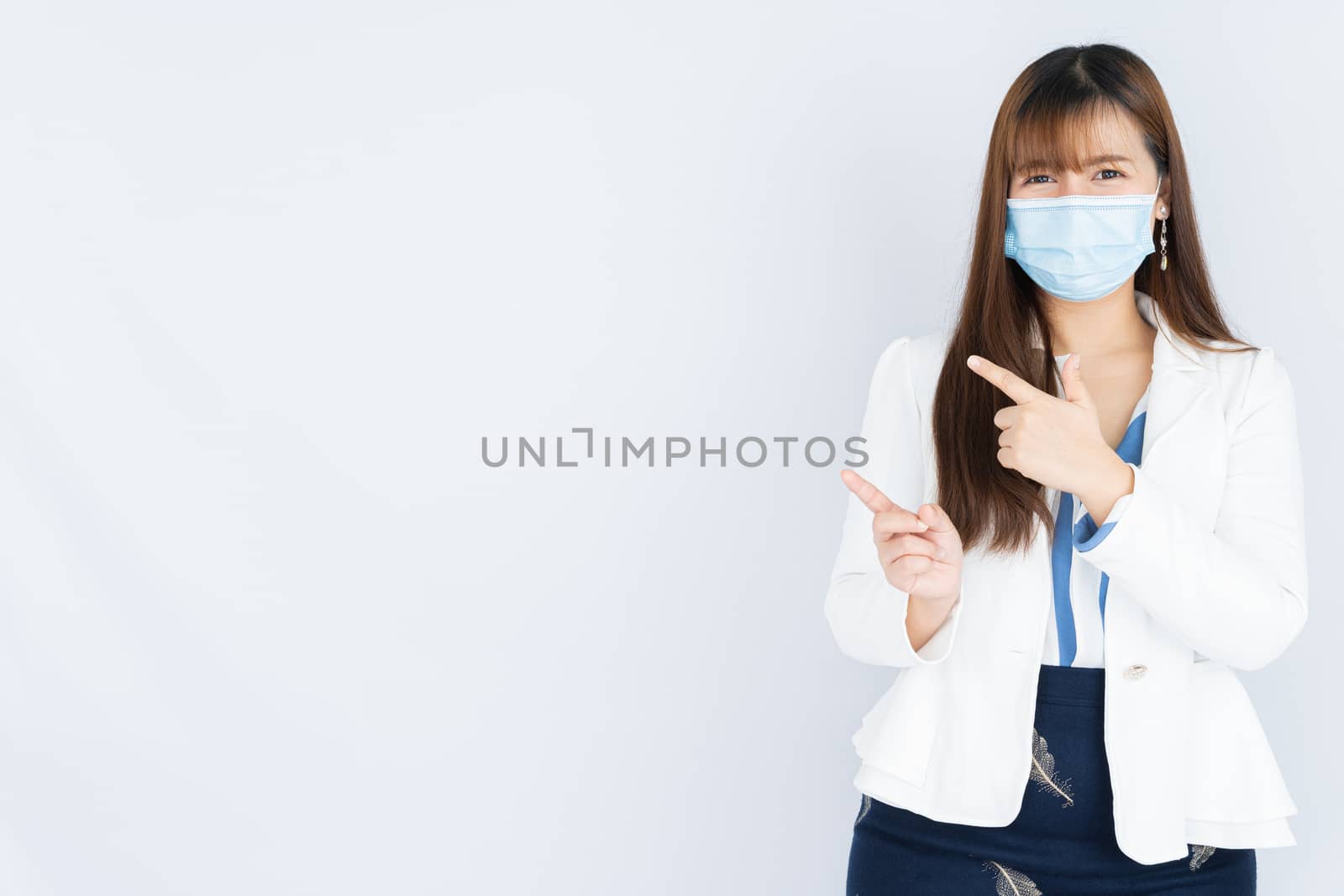 Smiling Asian business woman wearing a medical face mask and pointing hand to side blank space over grey background. Back to the normal concept.