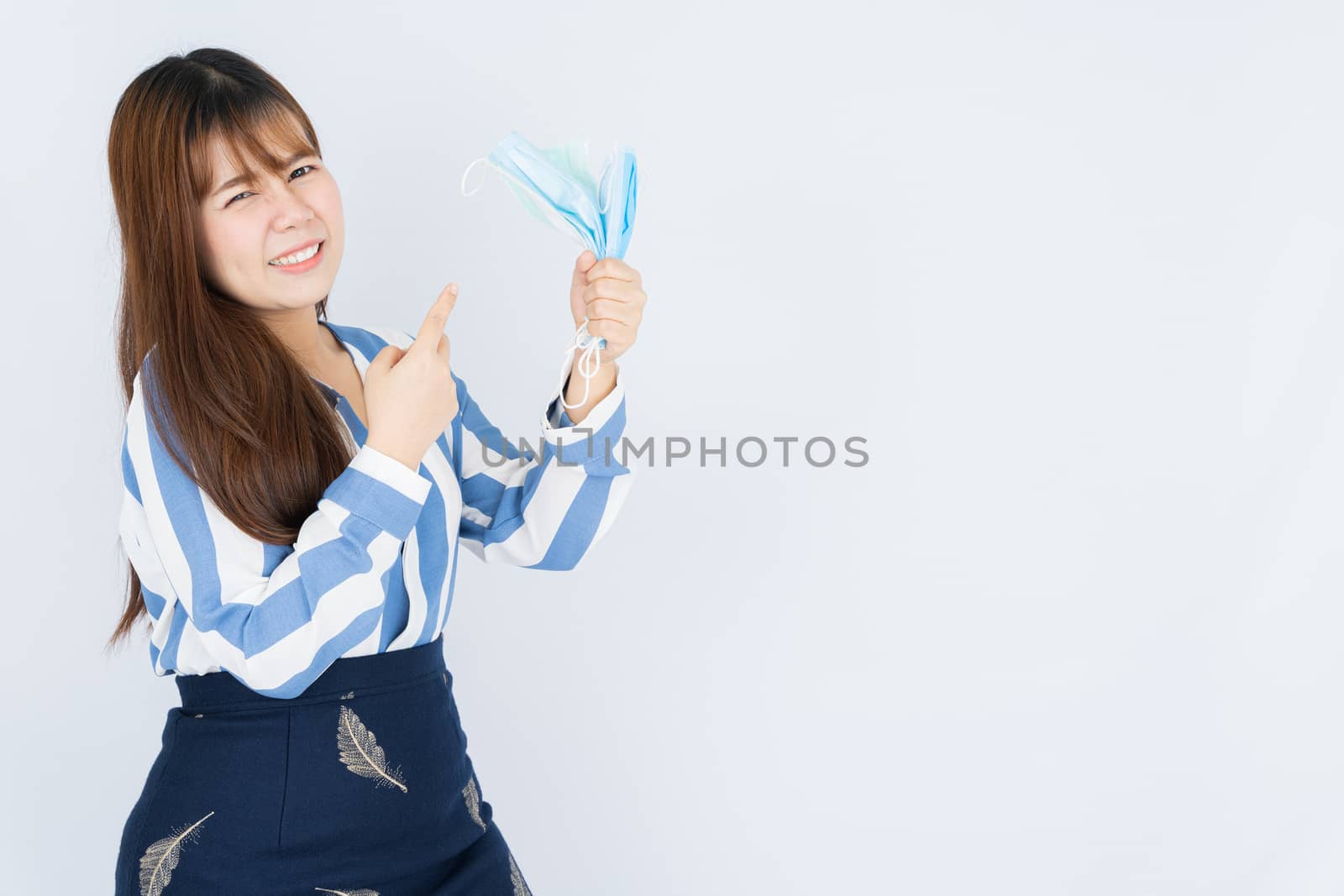 Smiling Asian business woman pointing finger to hand grasping mask over grey background. Back to the normal concept.